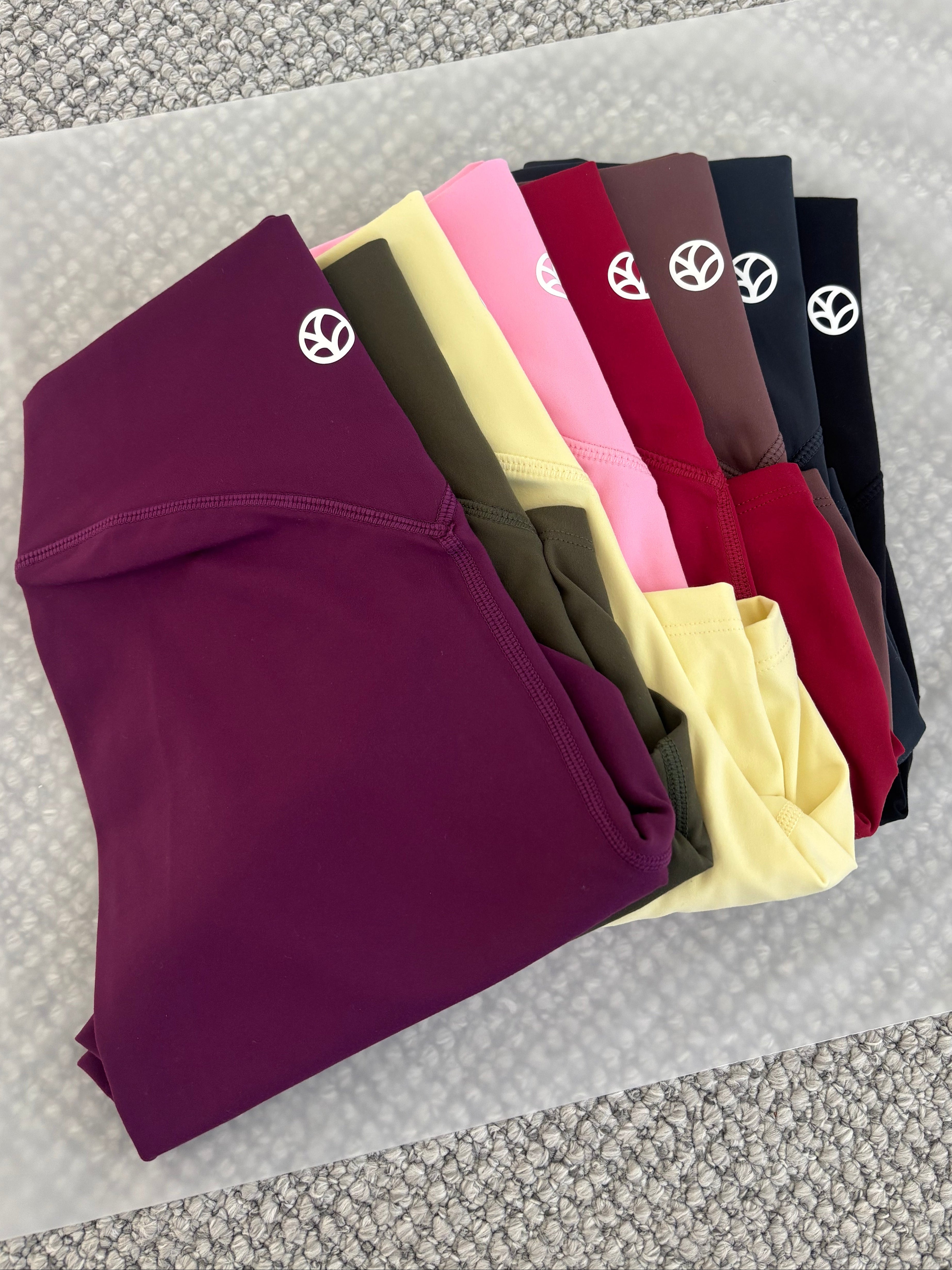 A fan arrangement of folded leggings in multiple colors, including purple, olive green, yellow, pink, red, brown, navy, and black, each featuring a small white circular logo on the upper leg, displayed on a white textured mat over a gray woven surface.
