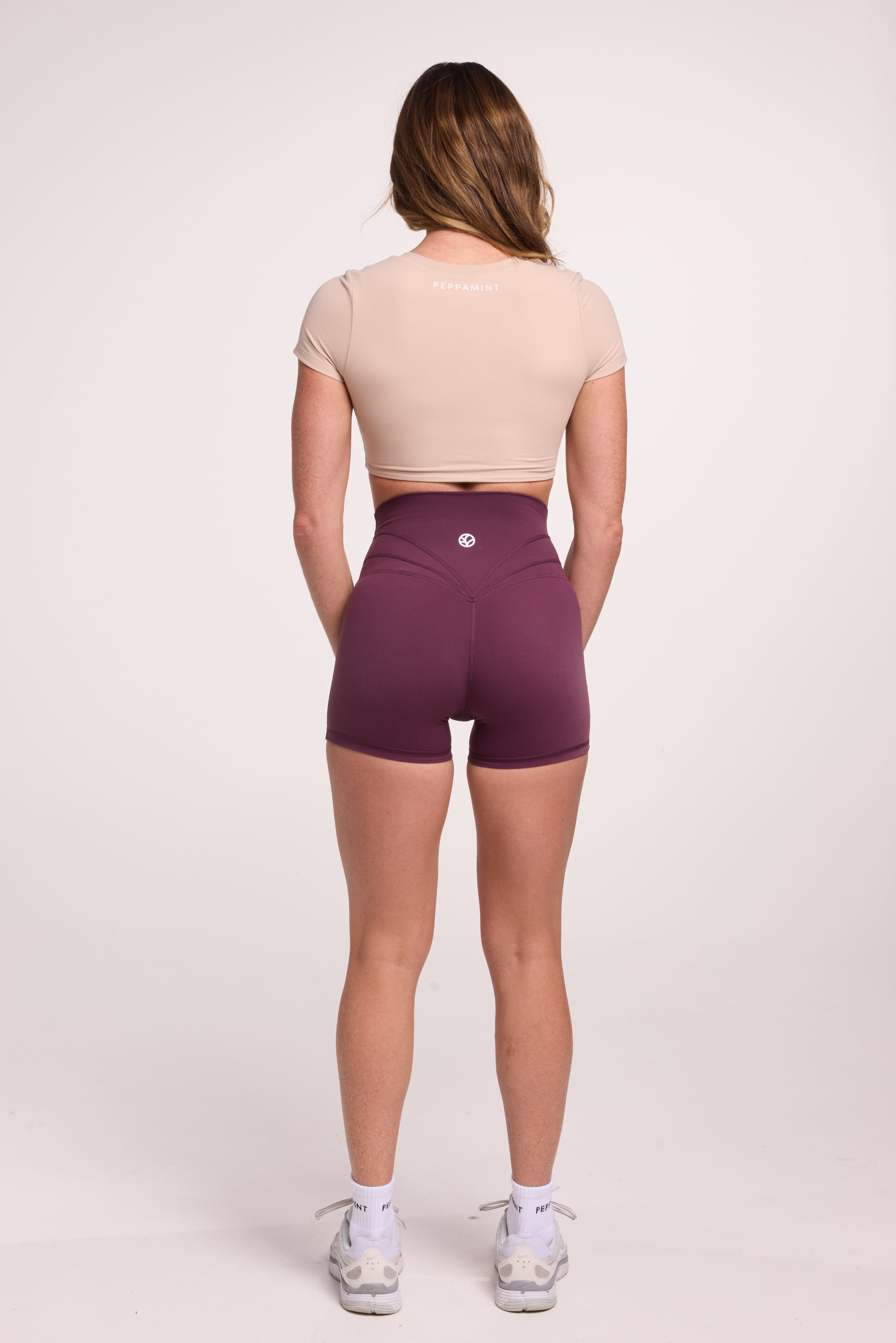 Buttery soft high waisted women's gym shorts with 4" inseam with curved stitched detailing for a butt shaping effect, and no front rise seam in plum purple paired with a V neck women's crop t-shirt with a supportive elasticated hem and triple layered fabric with removable pads for extra support in beige