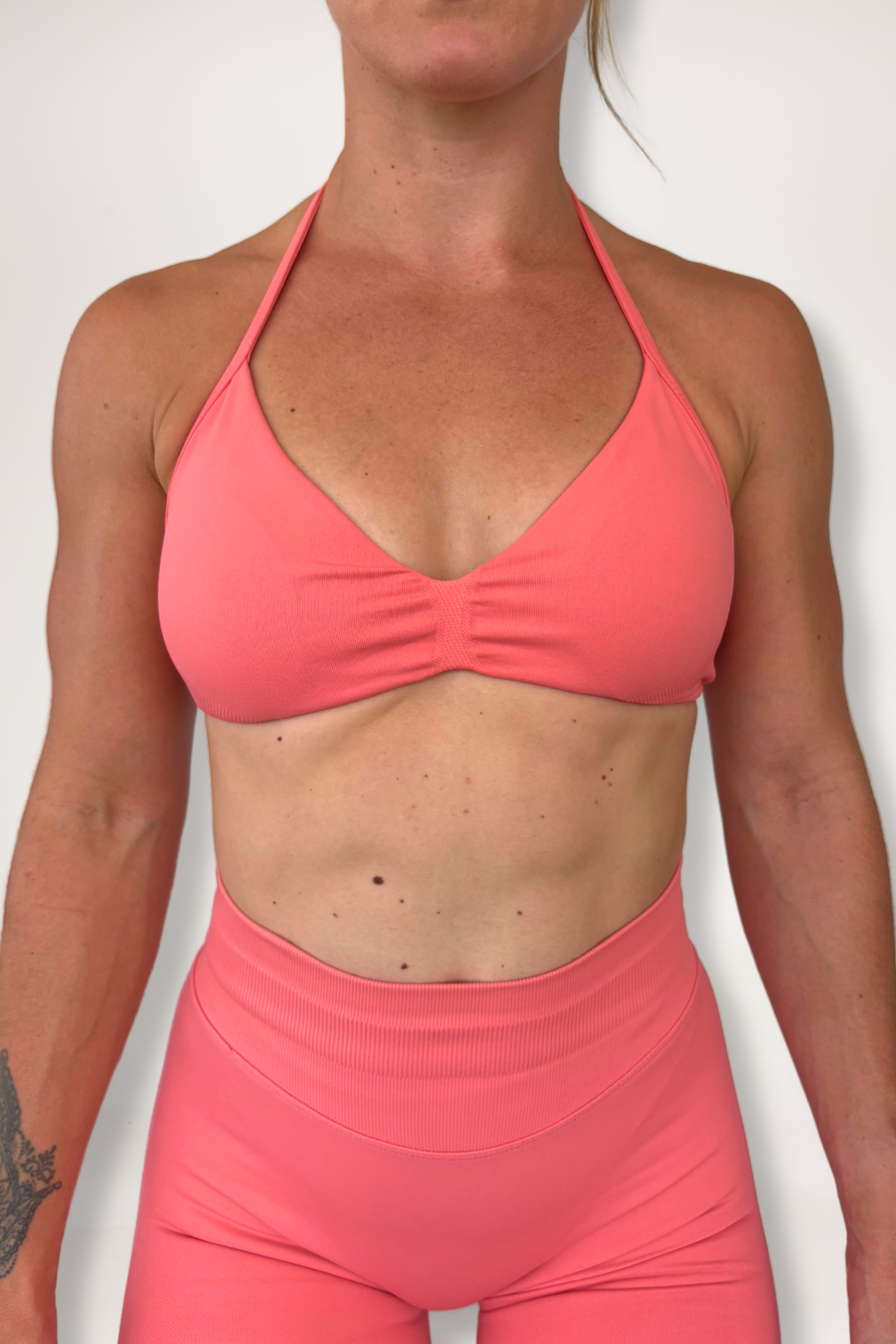 Minimalistic bra with breast contouring panels and a ruched front seam to effortlessy shape the breasts, with a sleek back straps and halter neck design for showcasing the back gains in coral
