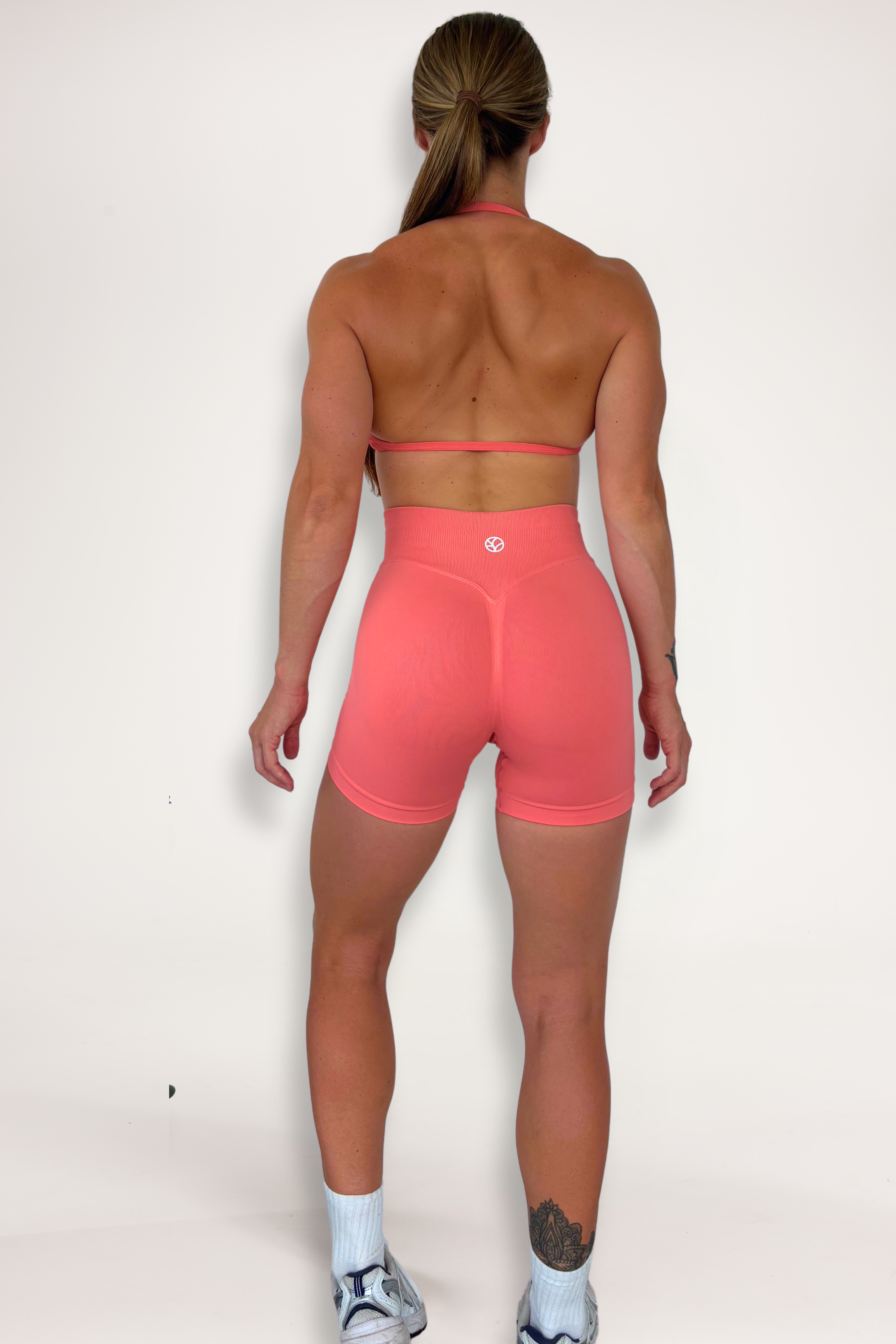 Seamless shorts with booty contouring panels, glute shaping seams, a thick supportive waistband paired with a minimalistic bra with sleek back straps and halter neck design for showcasing the back gains in coral