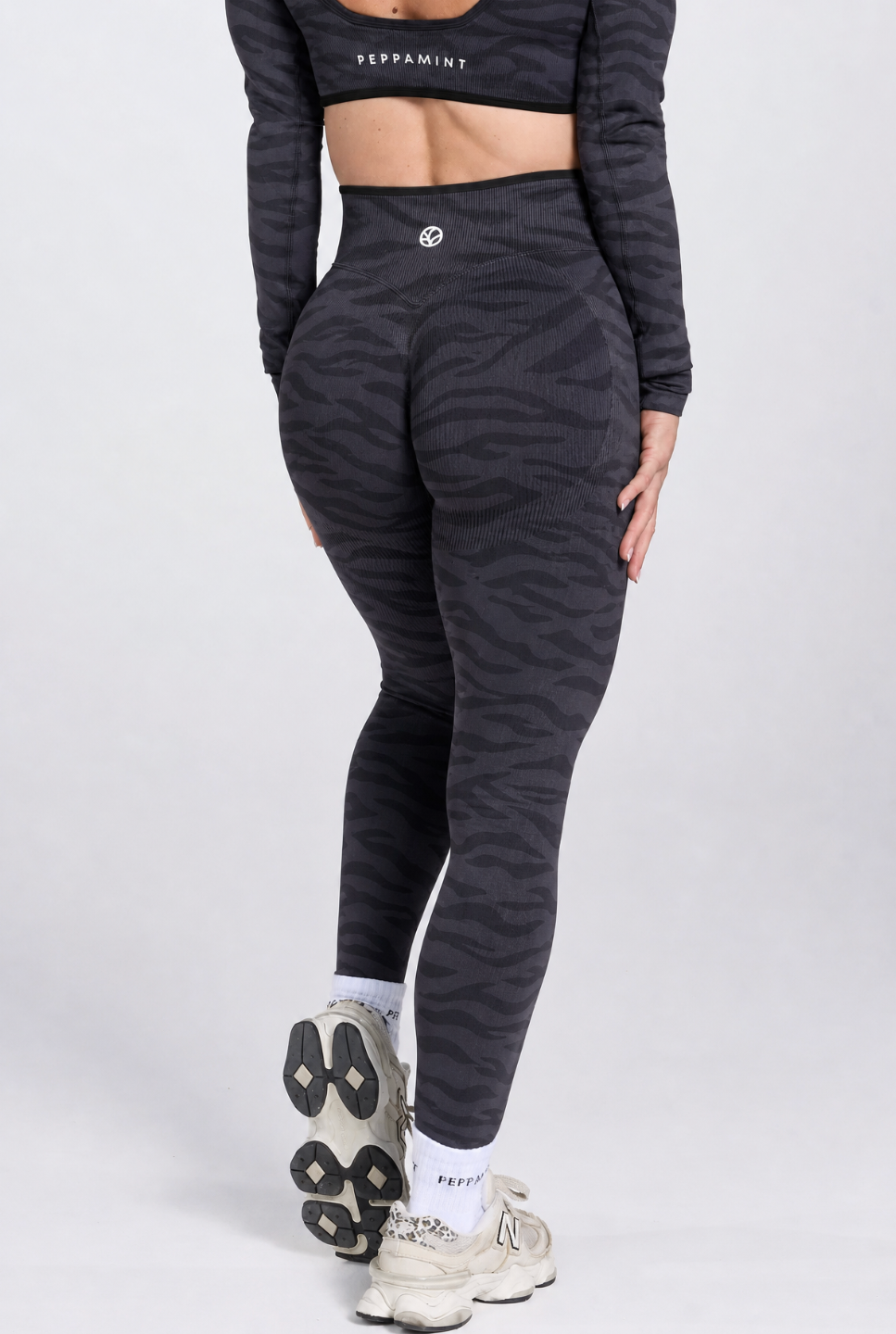 A model from the back wearing high-waisted booty shaping leggings with a dark grey and black zebra stripe pattern, a matching long-sleeve crop top, white socks, and beige sneakers on a light grey studio background.