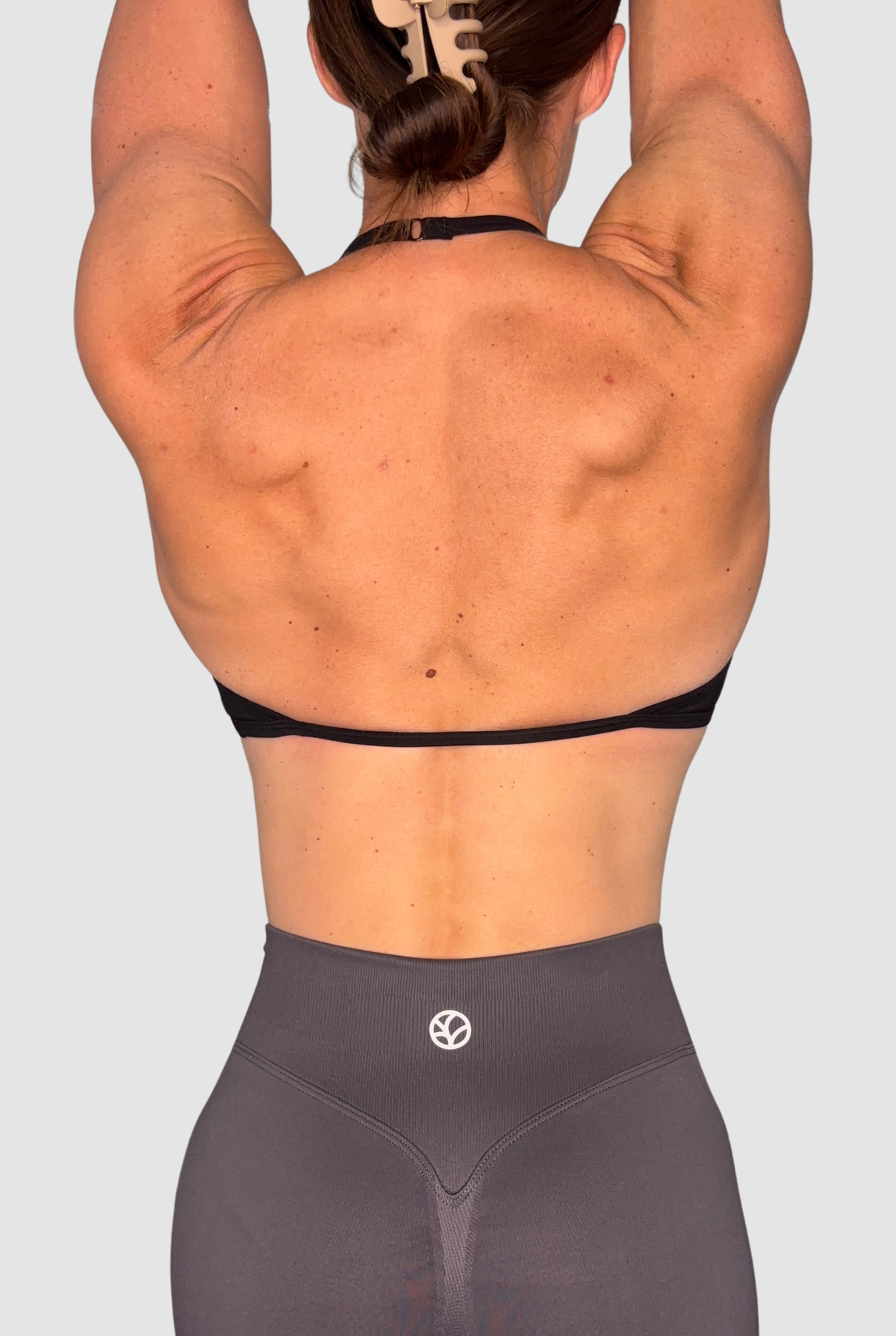 Rear view of a woman with her arms raised overhead, wearing a black sports bra and dark grey high-waisted shorts with a small white PEPPAMINT circular logo on the back waistband.