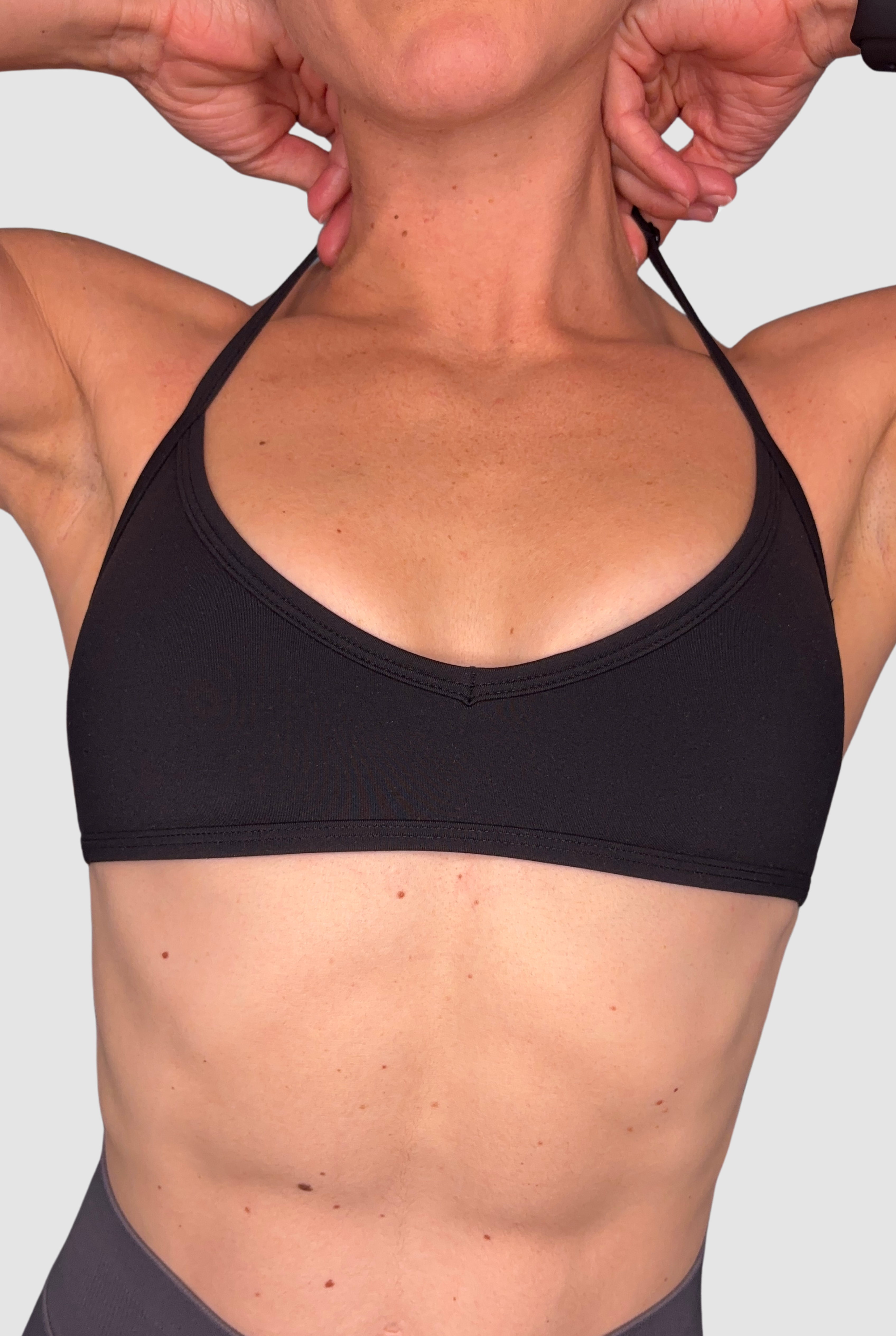 Close-up of a person wearing a black sports bra, with an adjustable halter neck, showing the front of the bra and upper torso against a light grey background.