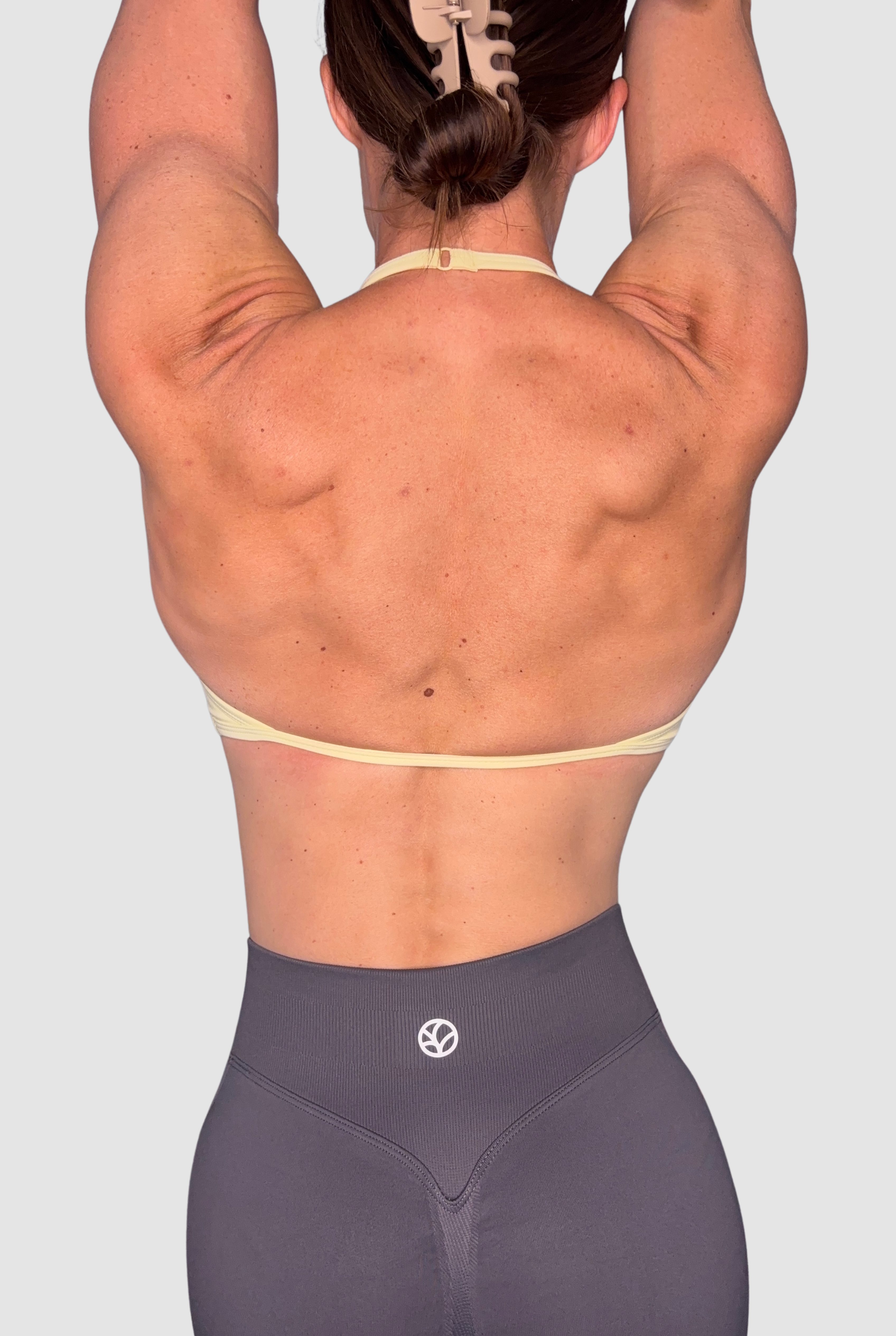 Rear view of a woman with her arms raised overhead, wearing a pale yellow sports bra and dark grey athletic leggings with a small white circular PEPPAMINT logo near the waistband, against a light grey background.