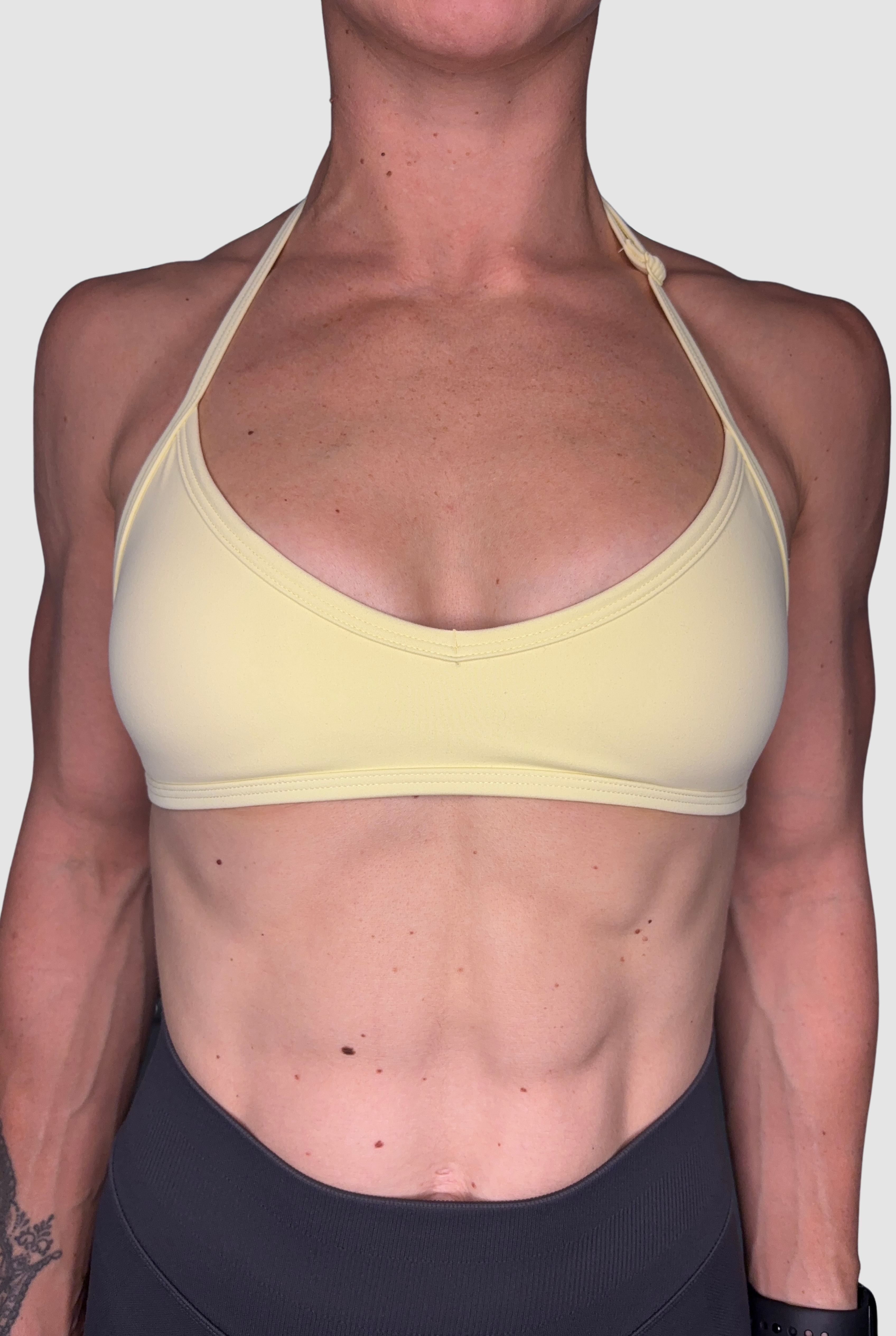 A close-up of a person wearing a pale yellow adjustable halter neck sports bra and dark grey athletic bottoms against a light grey background.