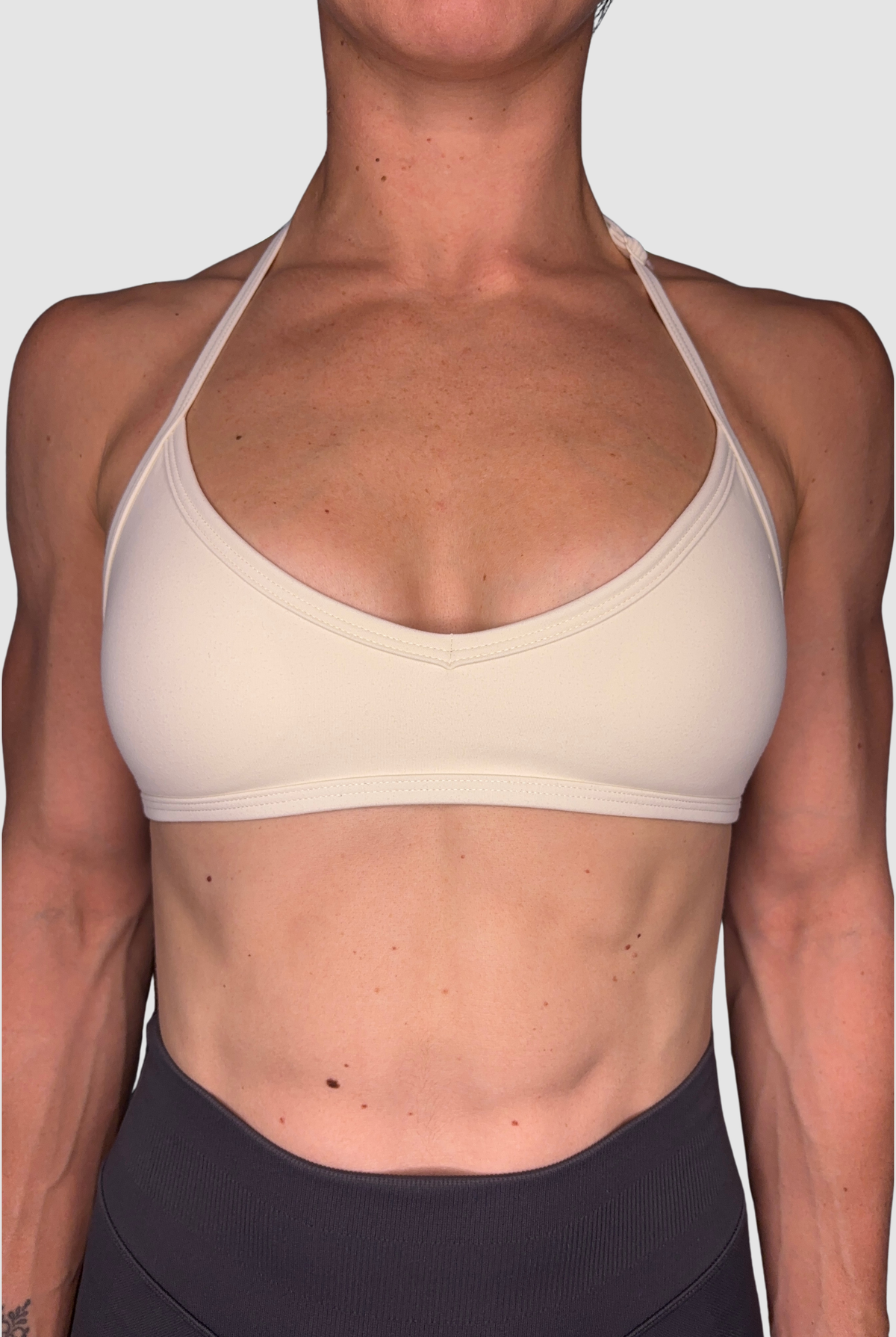 A close-up of a model wearing a light beige adjustable halter-neck sports bra and dark grey athletic bottoms against a light grey background.
