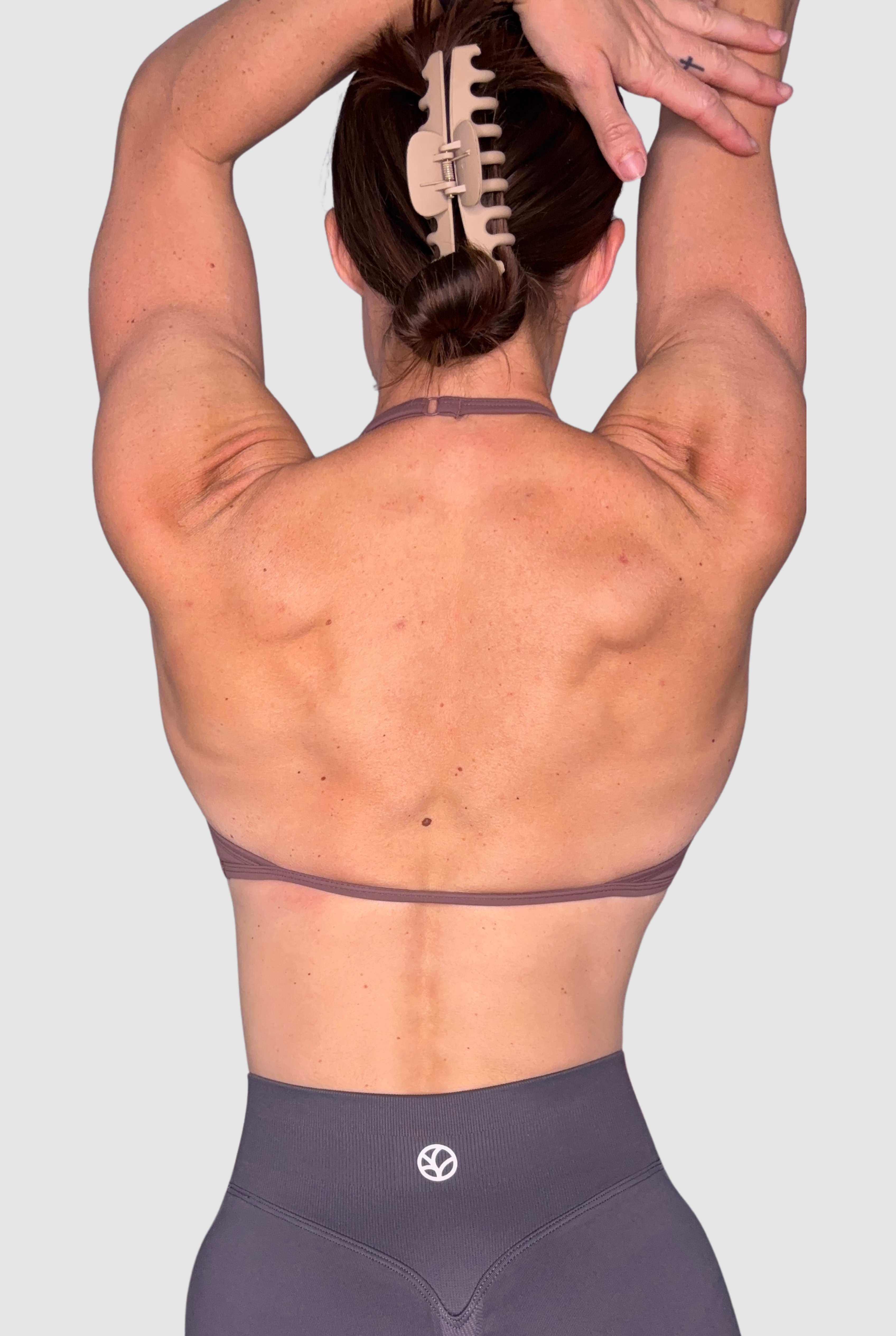 Rear view of a woman with her arms raised overhead, wearing a mauve-brown athletic sports bra and dark grey shorts with a small white PEPPAMINT logo at the centre back waistband.