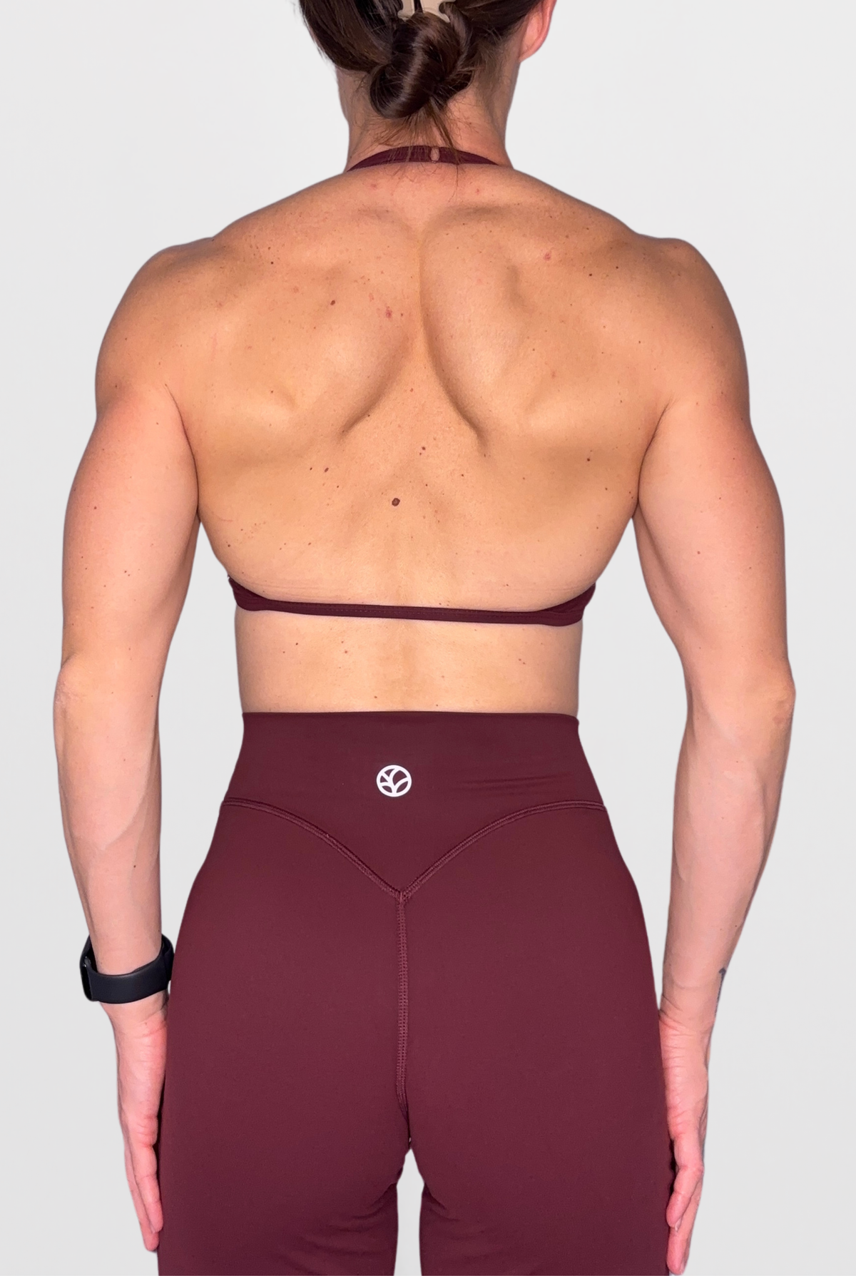 Rear view of a woman wearing a burgundy sports bra with a very thin back strap and adjustable halter neck and matching high-waisted leggings against a light grey background.