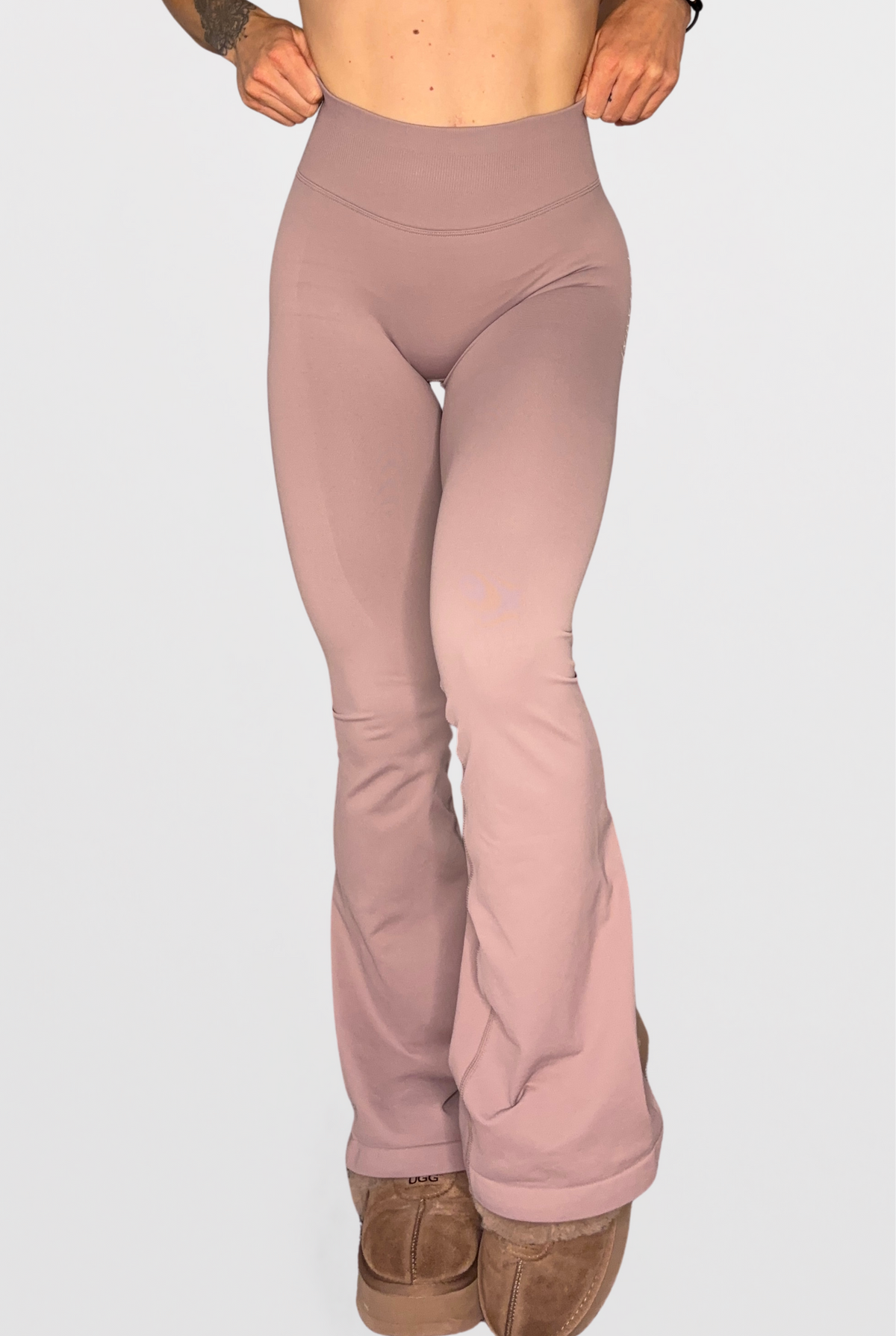 A person wearing stone high-waisted flared leggings and brown suede boots, shown from the waist down against a light grey studio background.