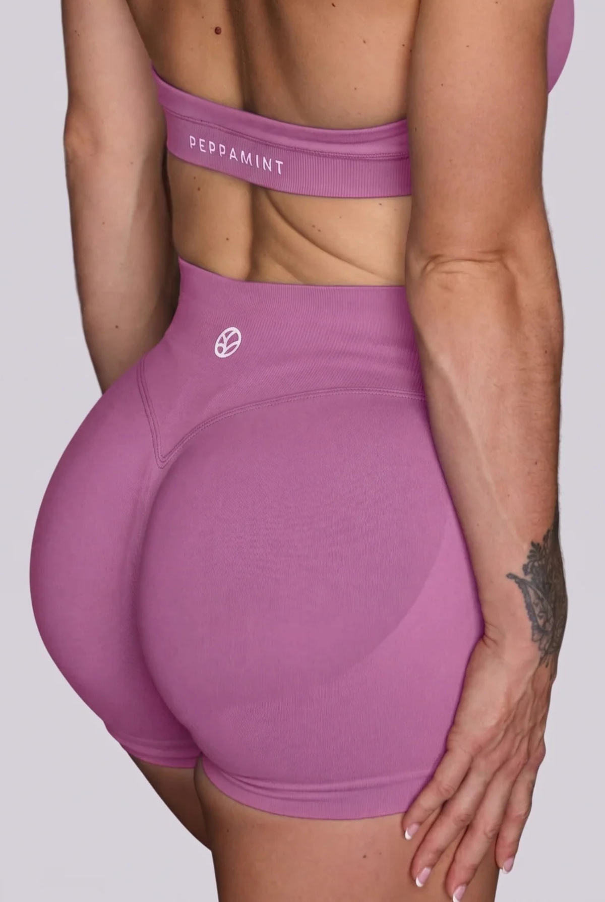 Close-up back view of a model wearing high-waisted athletic shorts and a matching sports bra in a dusty pink colour, with the brand name 'PEPPAMINT' visible on the sports bra band.