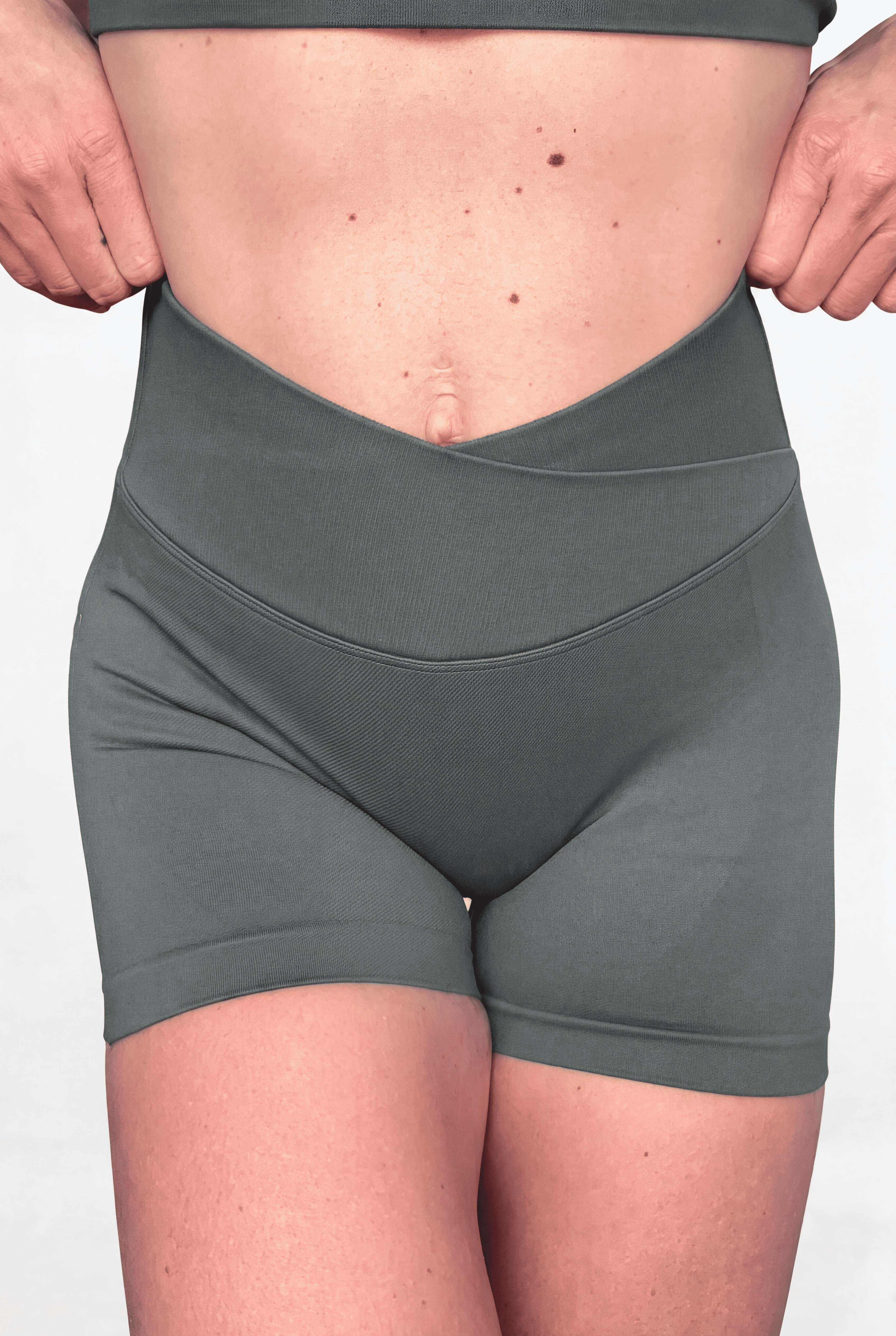 A close-up of a person wearing high-waisted athletic shorts with V crossover waistband in a sage green colour, holding the sides of the waistband to show its stretch and shape.