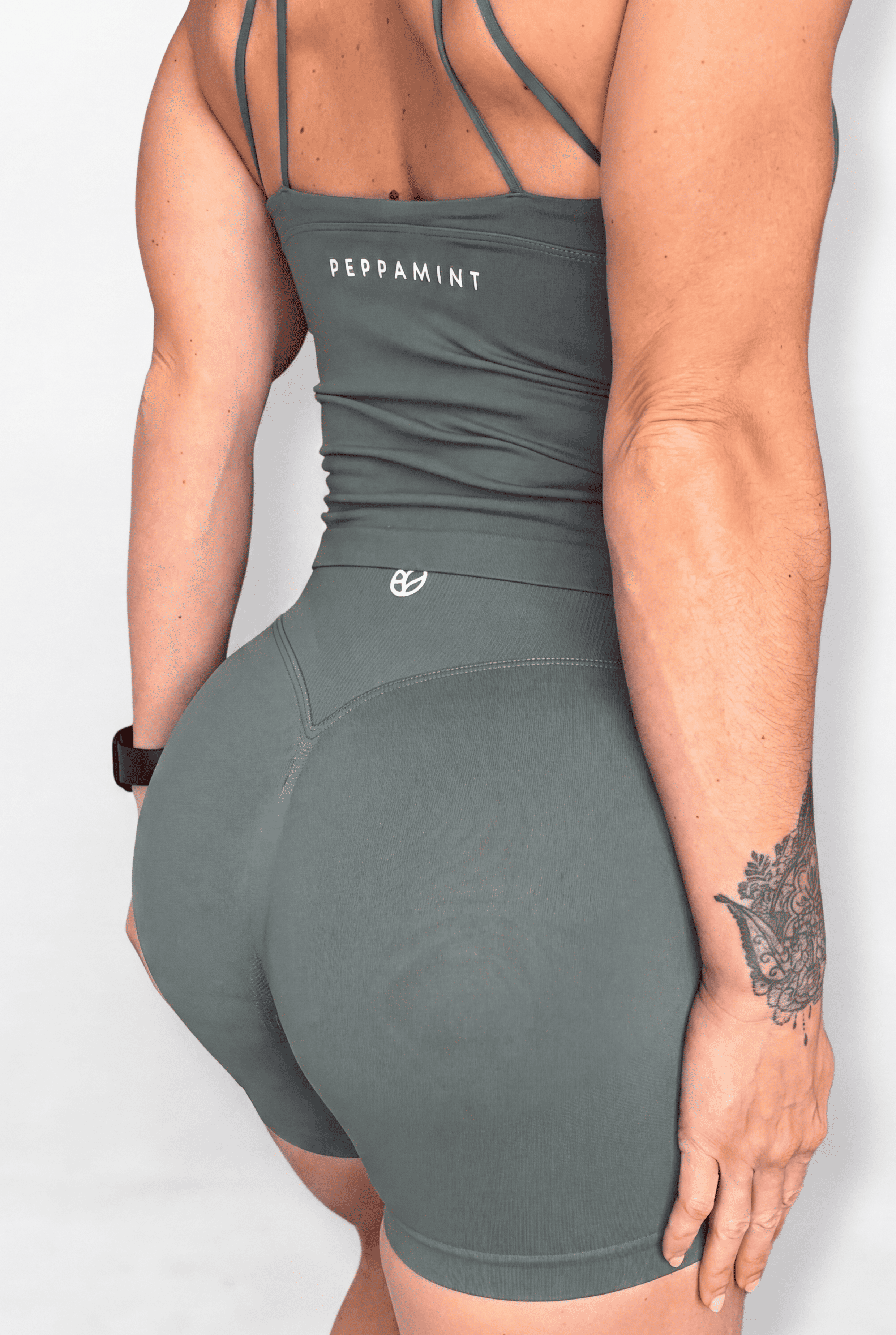 Rear view of a woman wearing a sage green athletic tank top with strappy back details and high-waisted shorts, featuring the brand name 'PEPPAMINT' printed on the back.