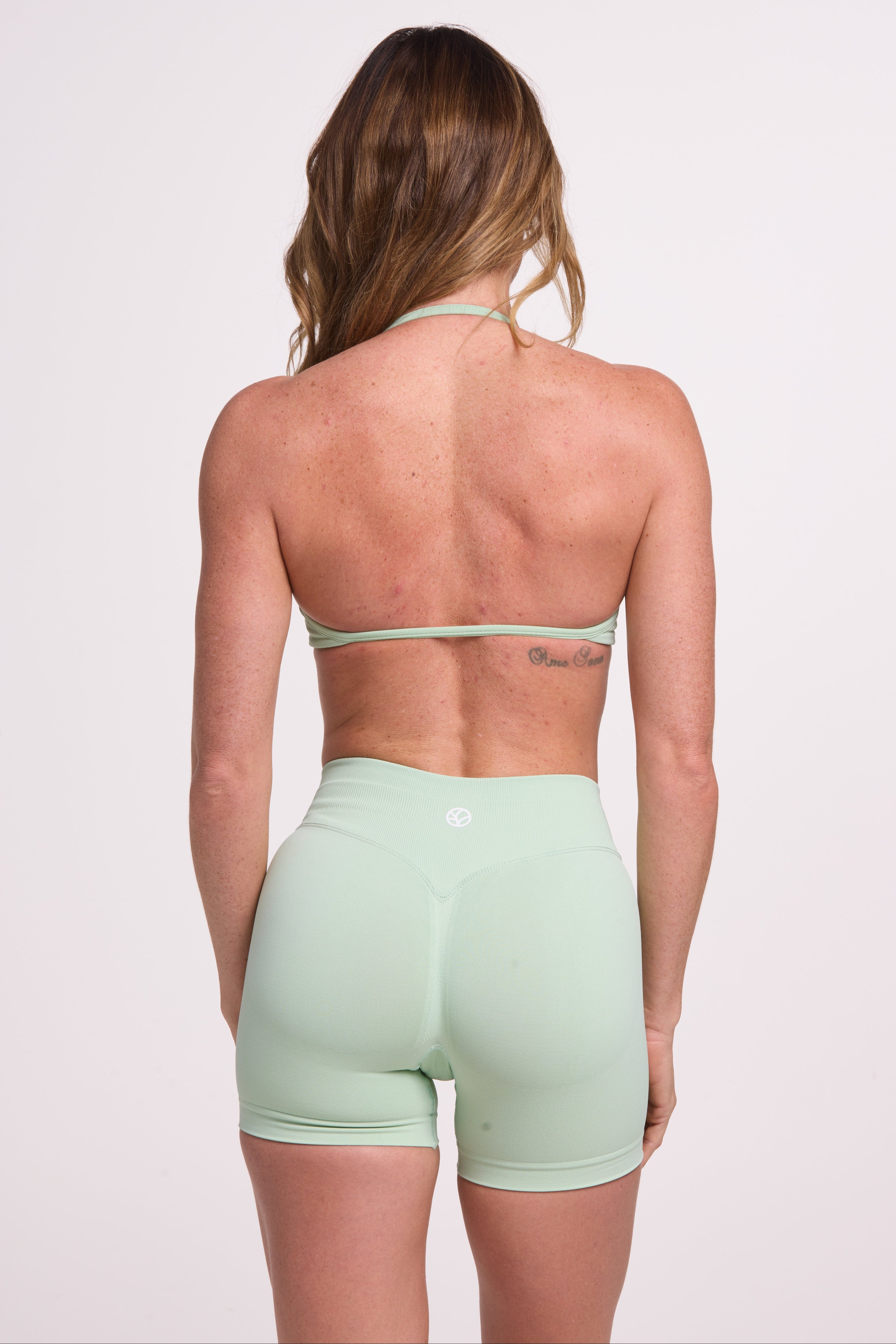 Minimalist bra with breast contouring panels and a ruched front seam to effortlessly shape the breasts, with a sleek back straps and halter neck design and seamless gym shorts with booty contouring panels, invisible scrunch technology, sweetheart shaped back seams and thick supportive fabric in mist green colour