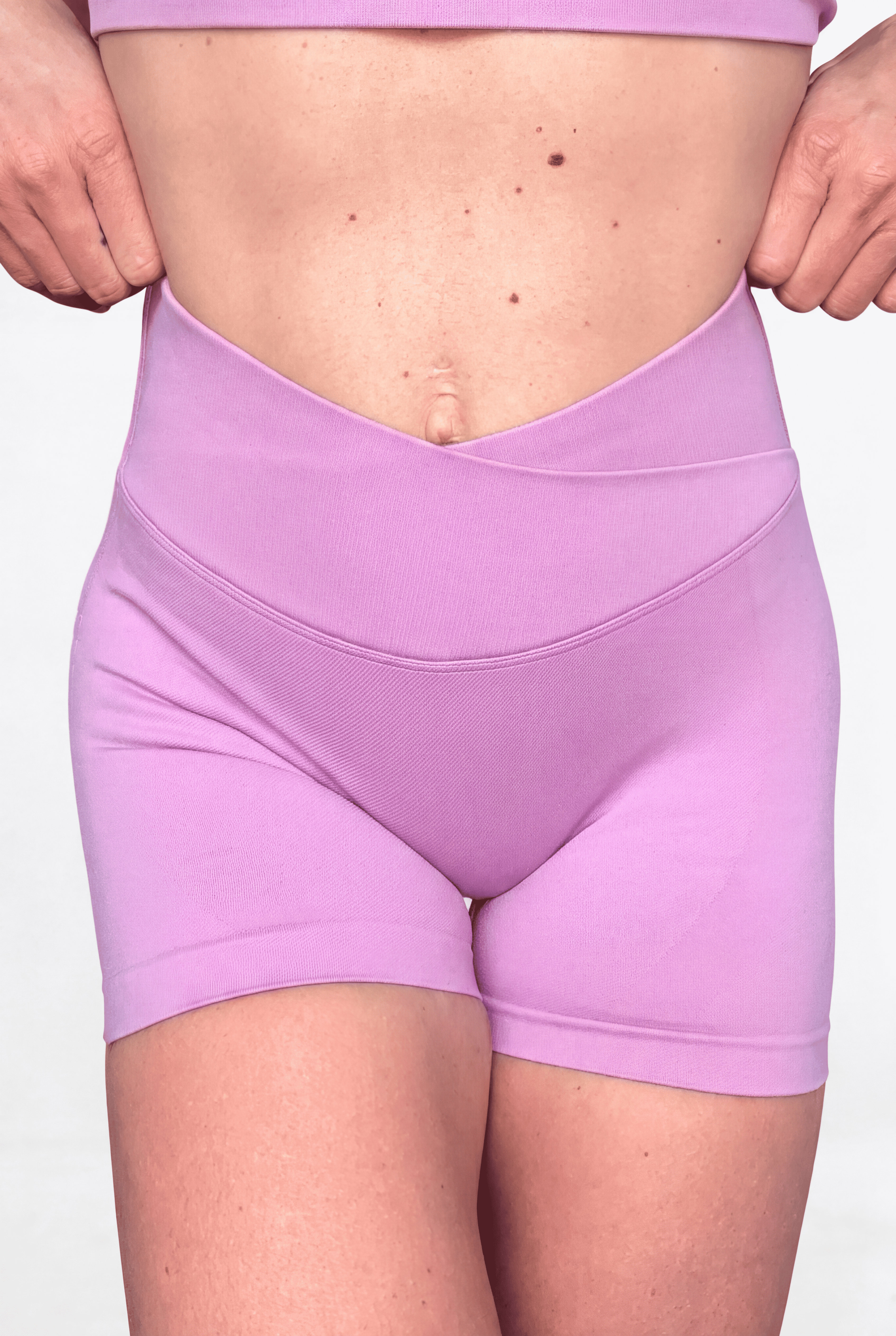 A close-up of a model wearing high-waisted, crossover-front athletic shorts in a solid light pink colour, shown from the waist to the upper thighs against a plain white background.