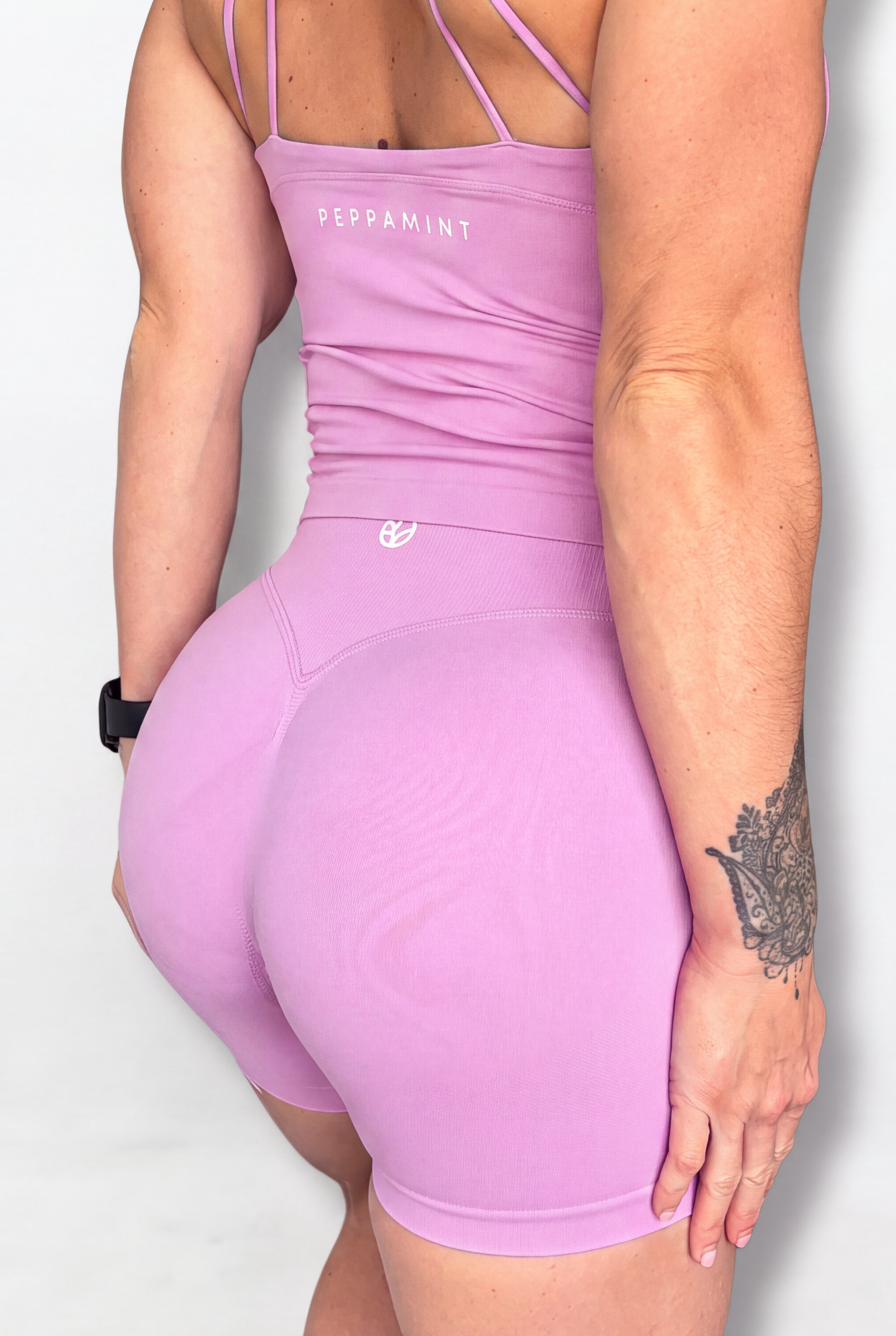 A woman shown from the back wearing a light pink tank with thin strappy back details and matching fitted shorts. The bodysuit has 'PEPPAMINT' printed in white near the upper back, and a small white logo on the waistband.