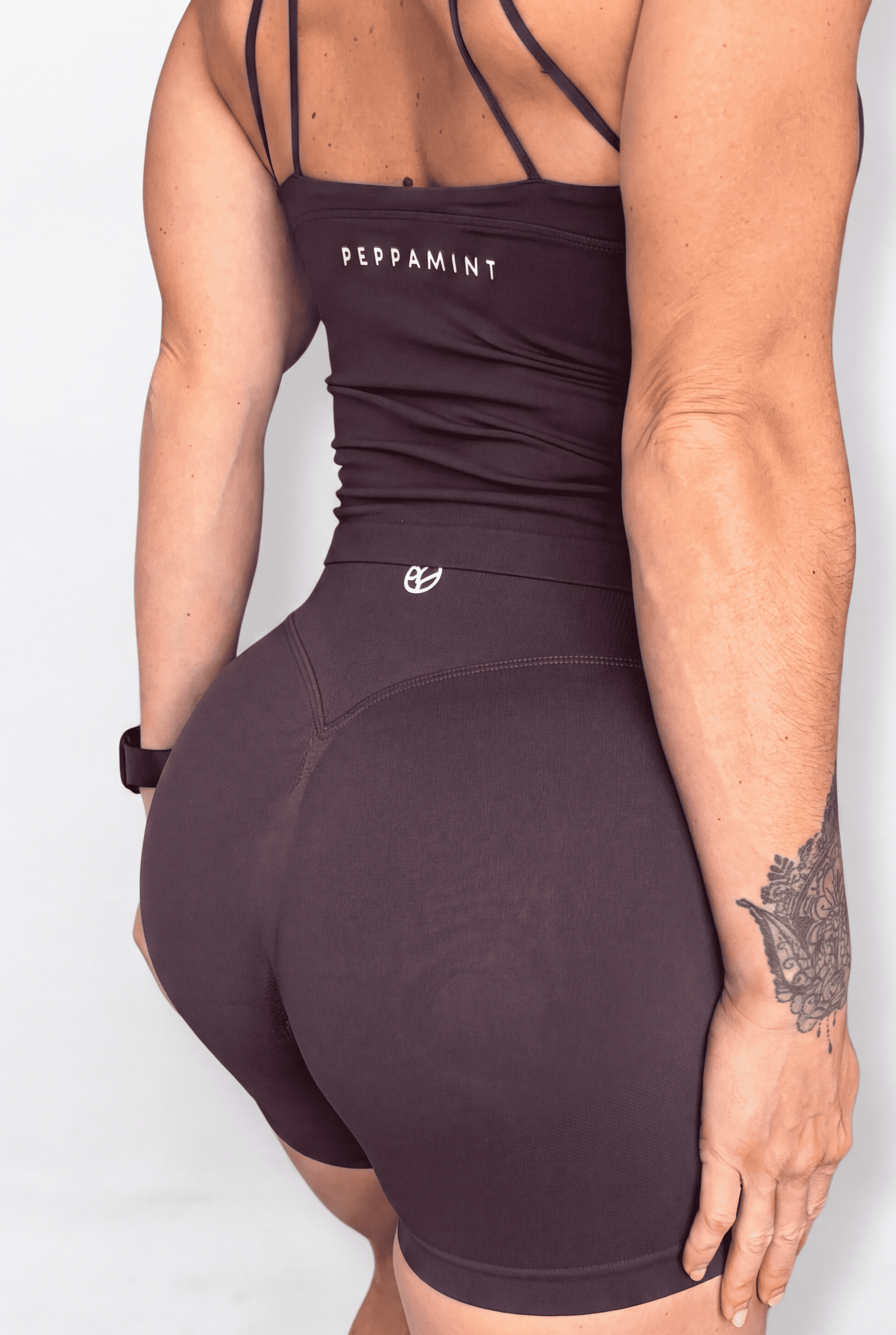 Rear view of a woman wearing a dark brown athletic tank with strappy back details and matching high-waisted shorts, on a light gray studio background.