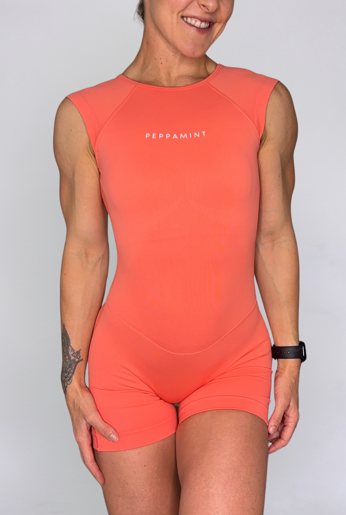 A woman wearing a sleeveless coral-orange athletic one-piece with matching shorts, featuring the word 'PEPPAMINT' printed in white on the chest, against a light grey studio background.