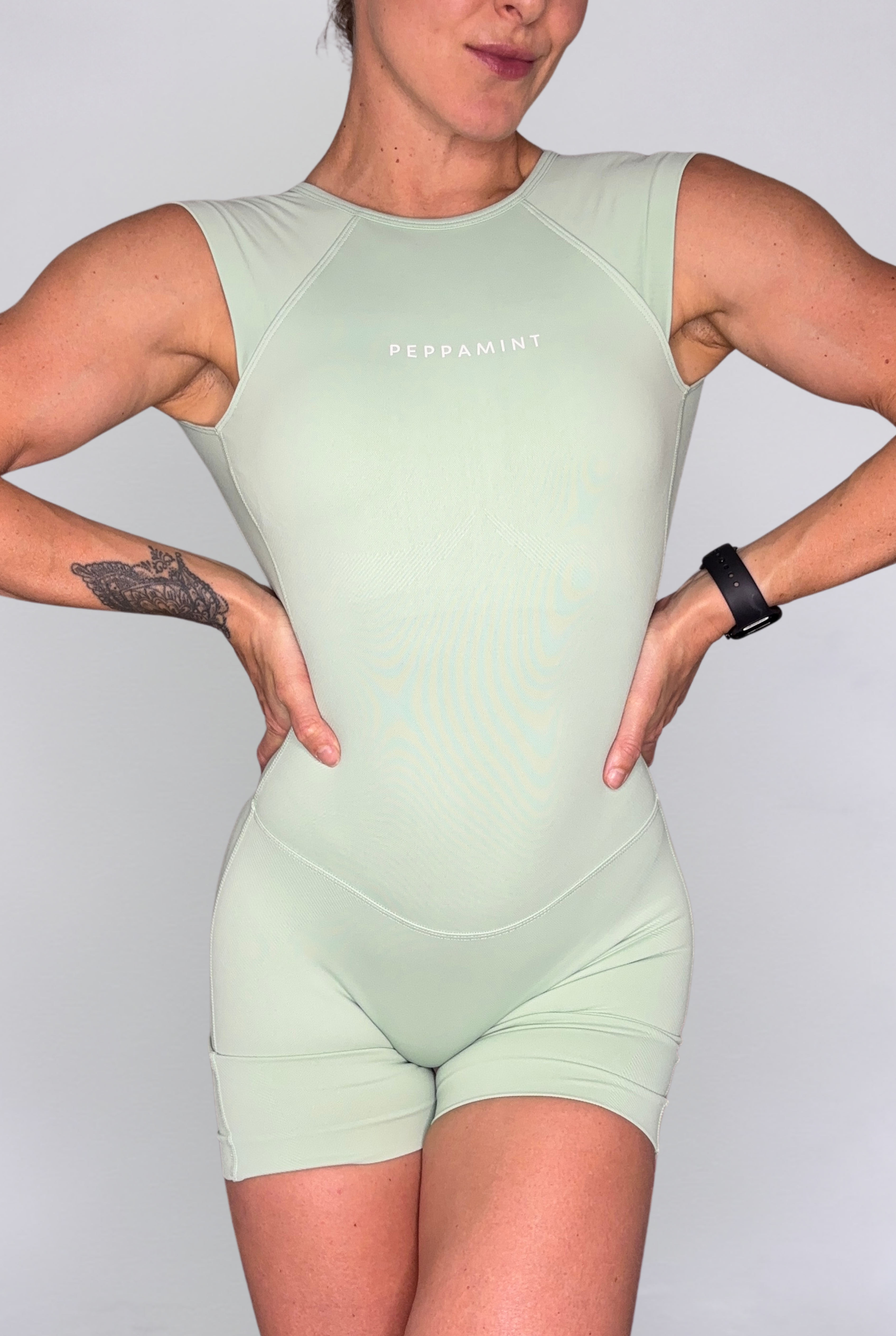 A woman modelilng a light mint green sleeveless athletic jumpsuit with 'PEPPAMINT' printed on the chest, set against a plain light grey background.