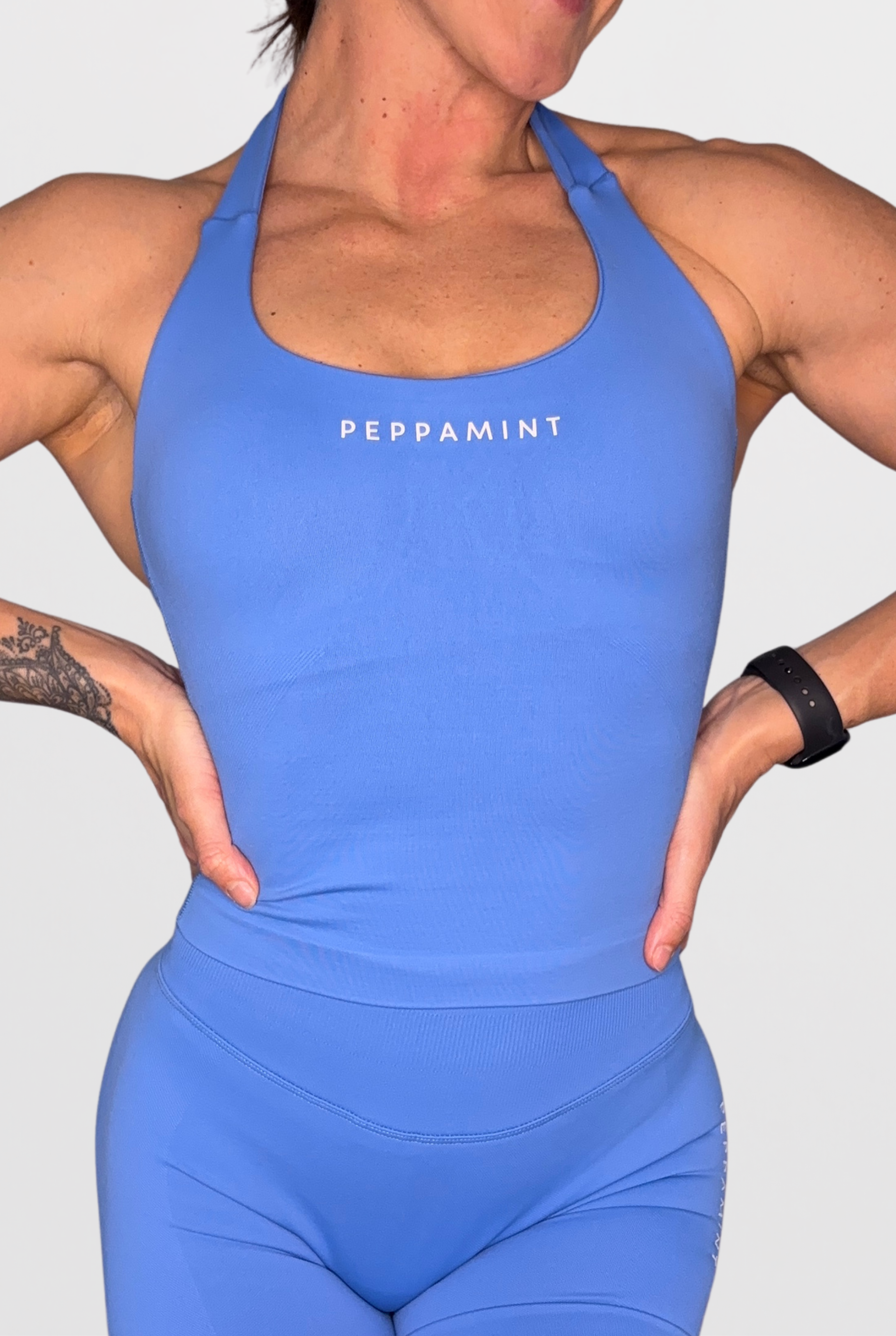 A close-up of a woman wearing a Powder Blue athletic tank top and matching leggings with the word 'PEPPAMINT' printed in white across the chest, against a light grey background.