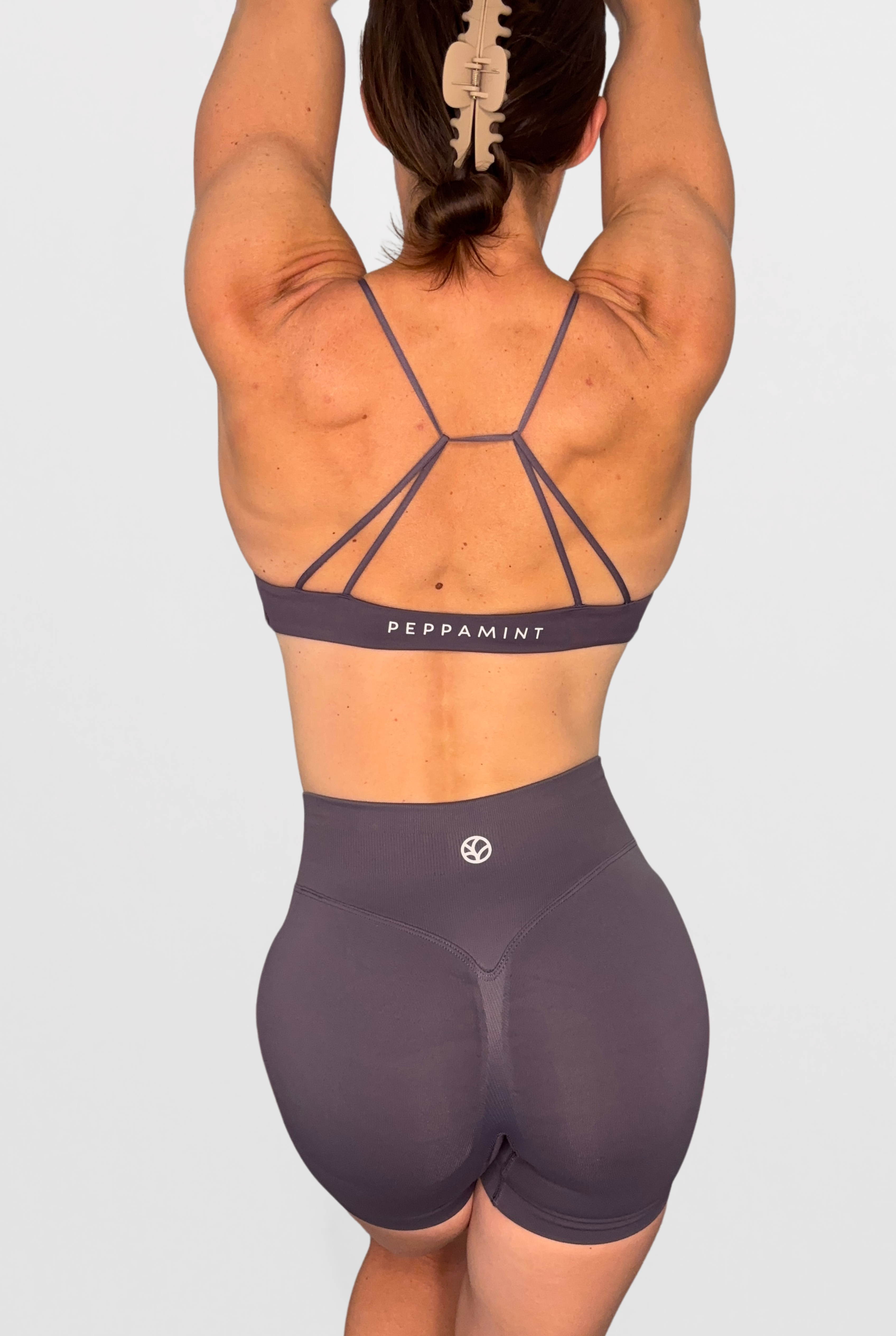 Rear view of a woman wearing a matching athletic set consisting of a strappy sports bra and high-waisted shorts in a dusty purple with the brand name 'PEPPAMINT' printed on the underband.