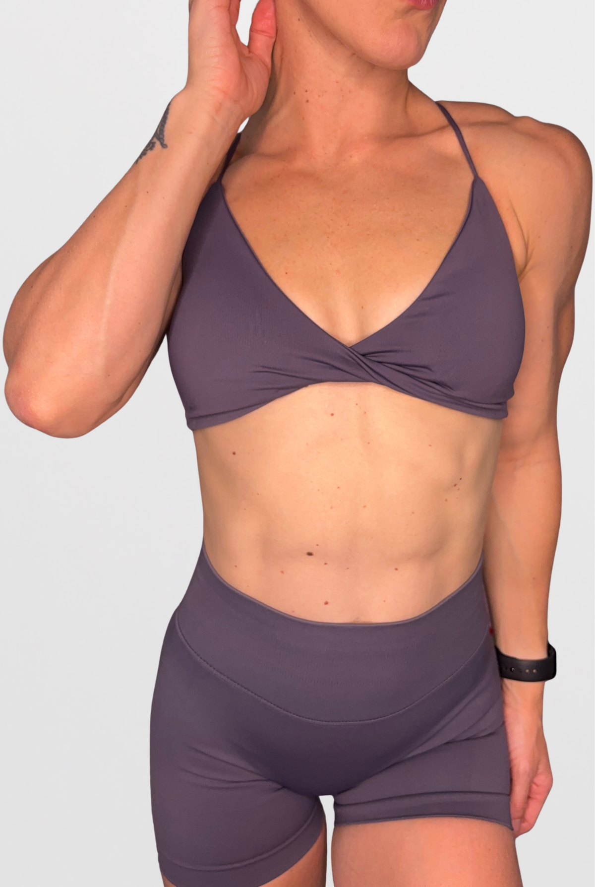 A woman wearing a matching two-piece athletic set consisting of a twisted-front sports bra and high-waisted shorts in a muted dusty purple colour, shown against a plain light grey background.