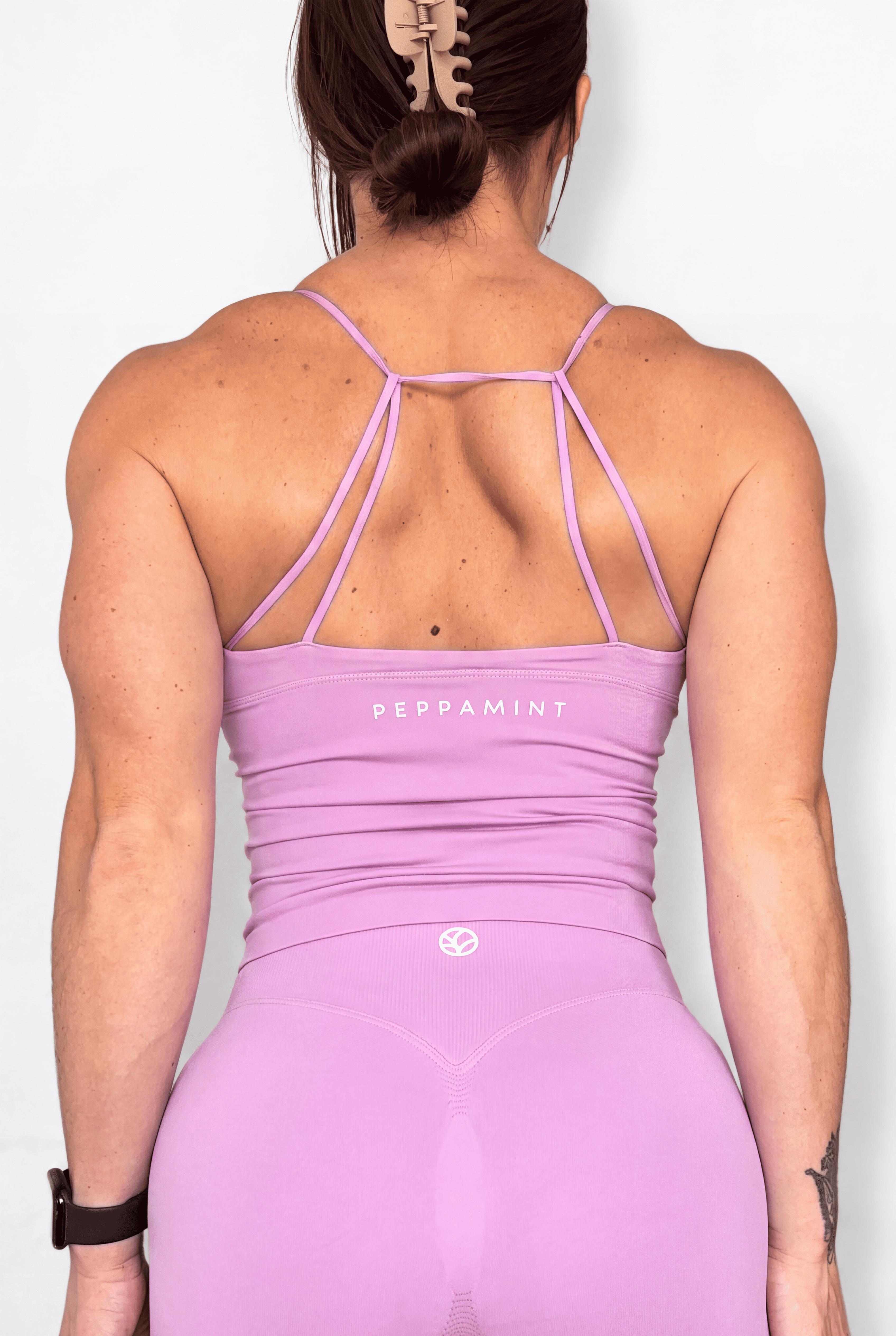 Rear view of a woman wearing a light pink athletic tank with thin crisscross back straps and the brand name 'PEPPAMINT' printed on the upper back, set against a plain white background.