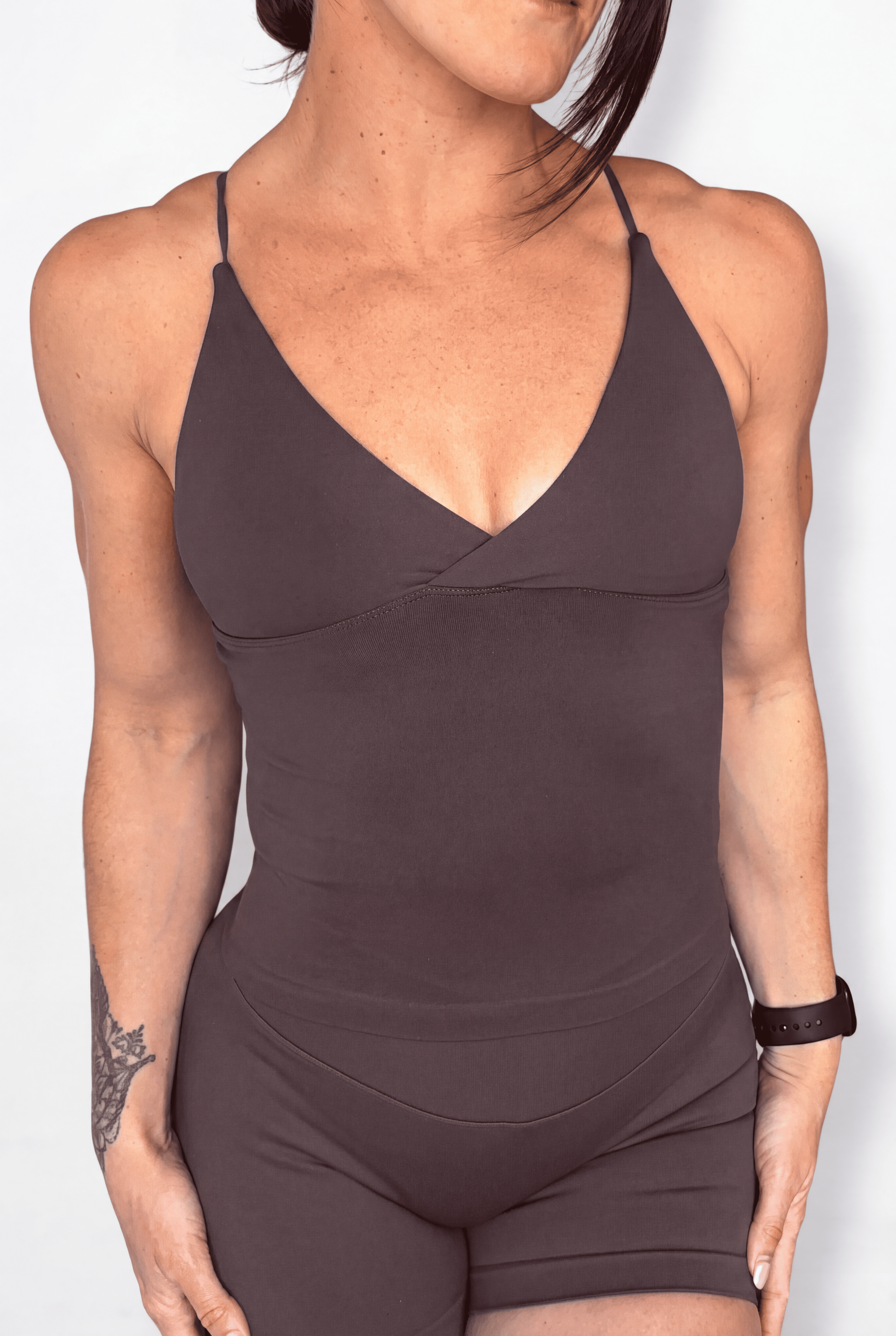 A woman wearing a dark brown tank top with thin spaghetti straps and matching shorts, shown from the neck down against a light gray background.