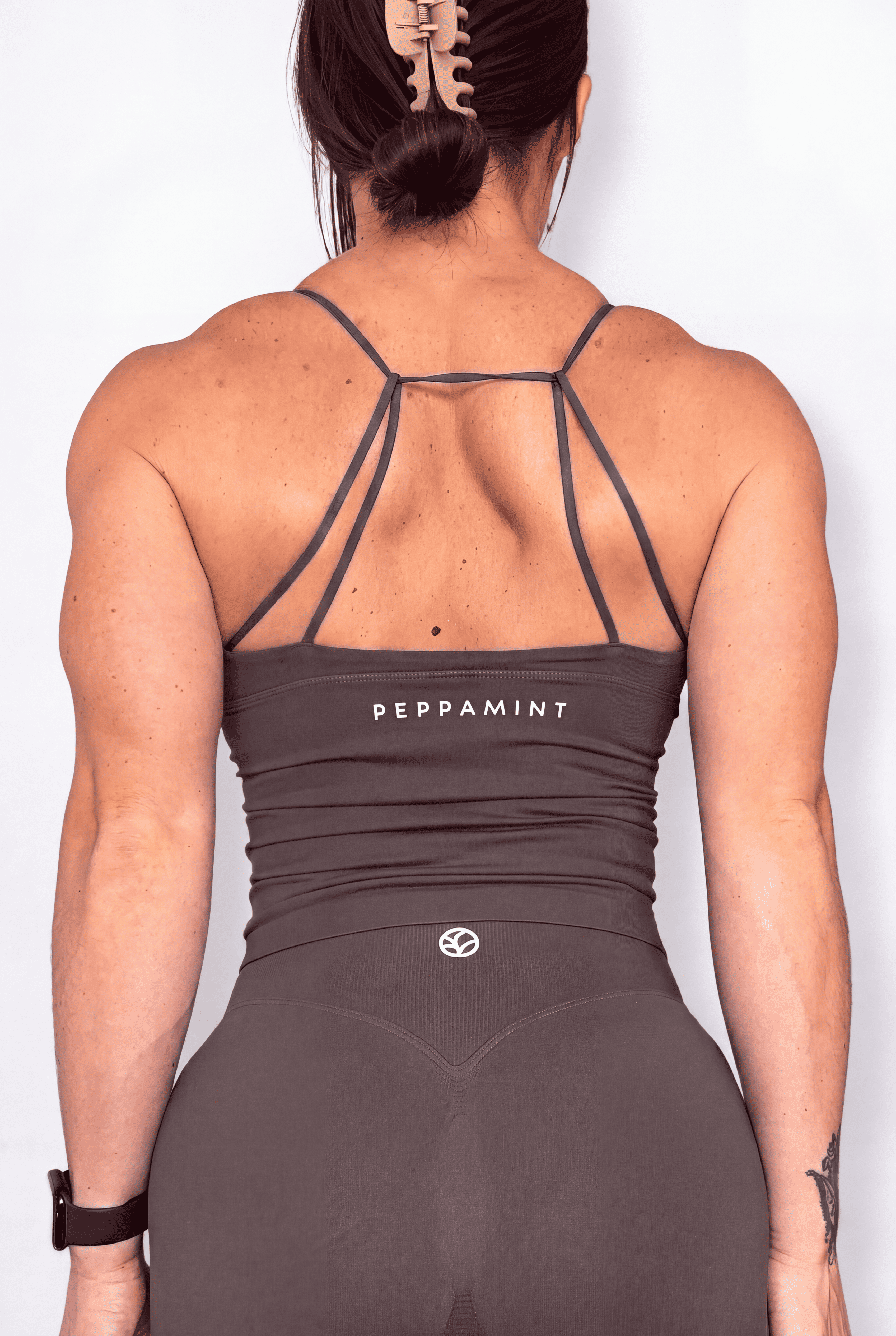 Rear view of a woman wearing a dark brown athletic tank top and matching short with the brand name 'PEPPAMINT' printed on the back, set against a plain white background.