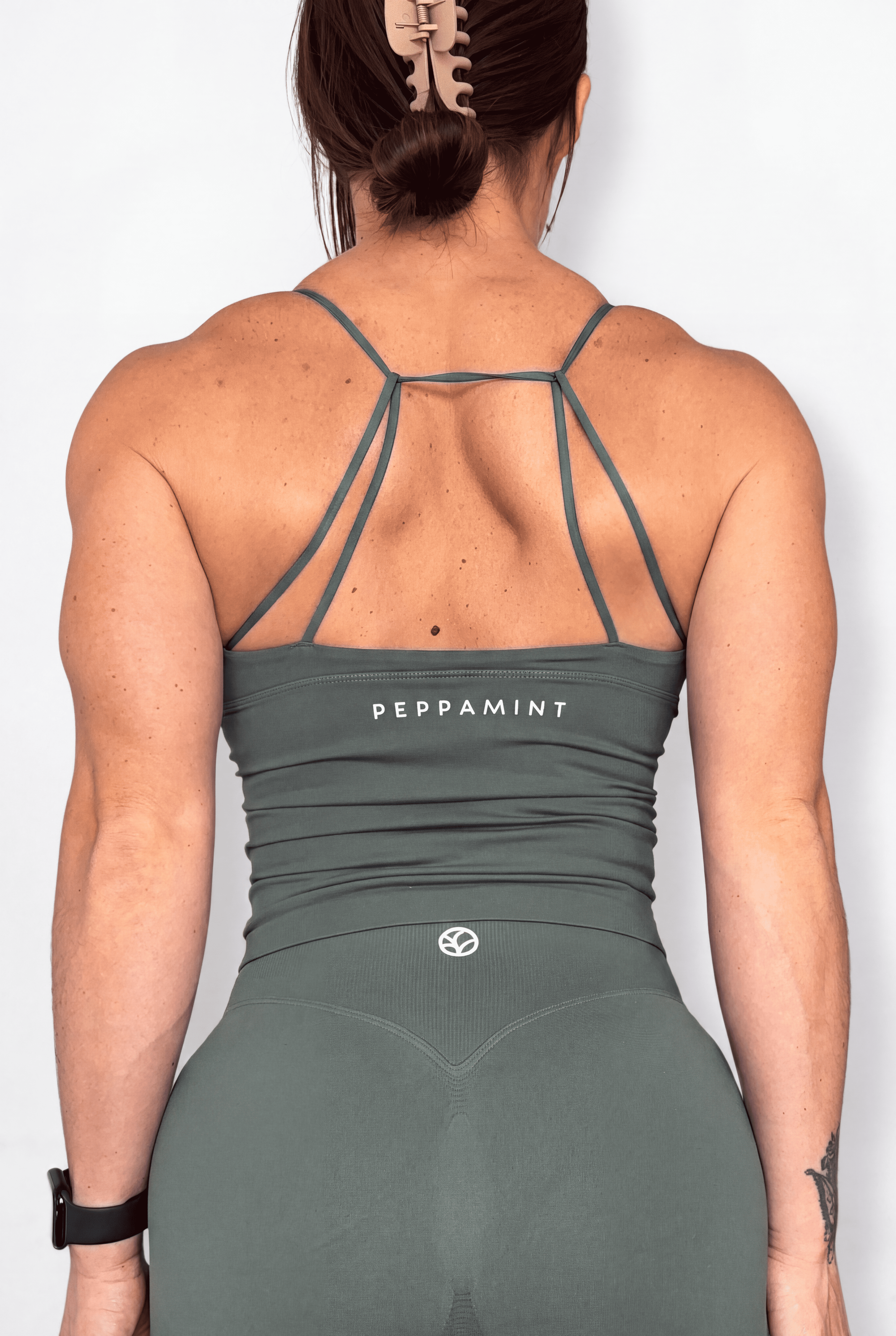 Rear view of a woman wearing a sage green athletic tank top with 'PEPPAMINT' printed in white on the back, set against a plain light gray background.