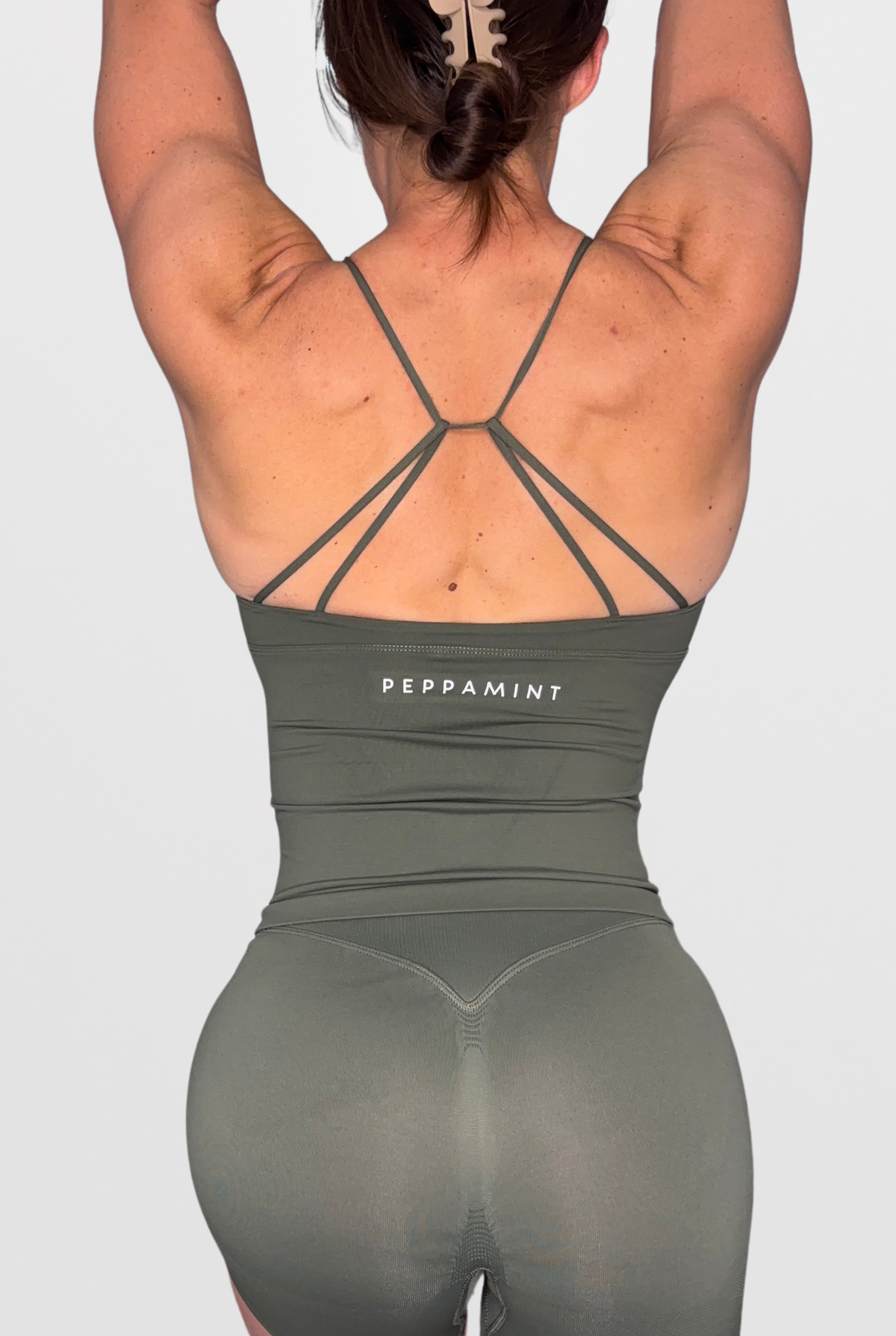 Rear view of a woman wearing an olive green athletic bodysuit with crisscross back straps and the brand name 'PEPPAMINT' printed in white on the back, against a light grey background.