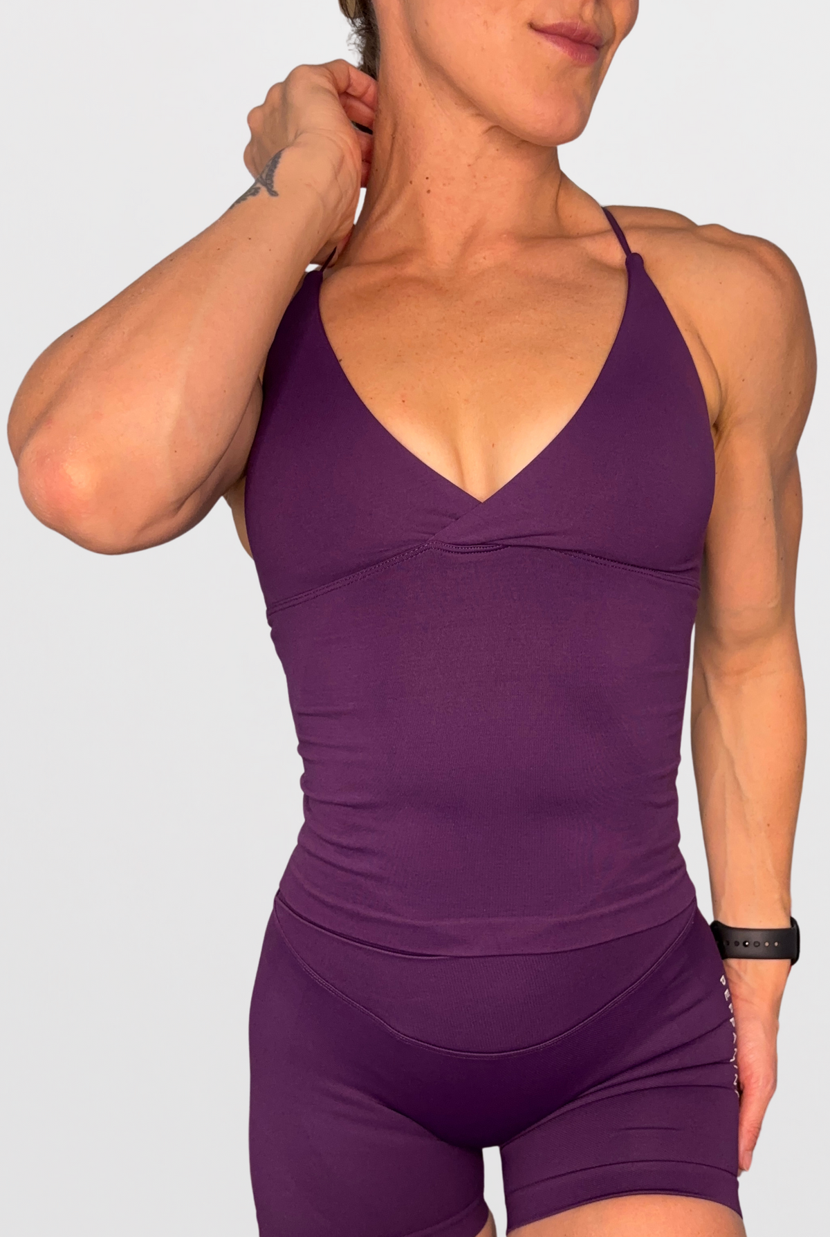 A woman wearing a matching grape purple athletic bodysuit with a deep V-neck and fitted shorts, shown against a light grey studio background.