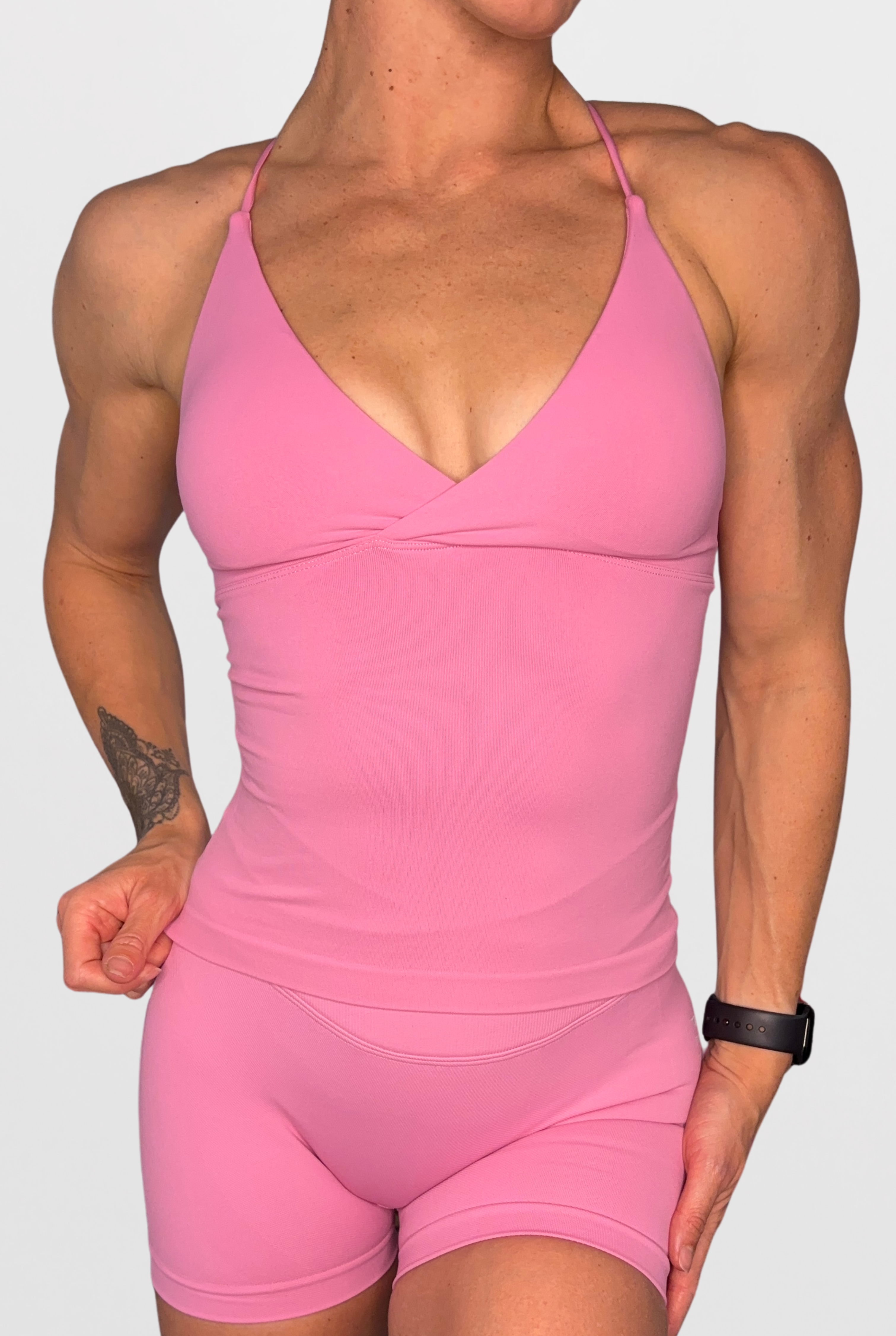 A woman wearing a rose pink athletic tank top with deep V neck and matching fitted shorts against a light grey studio background.