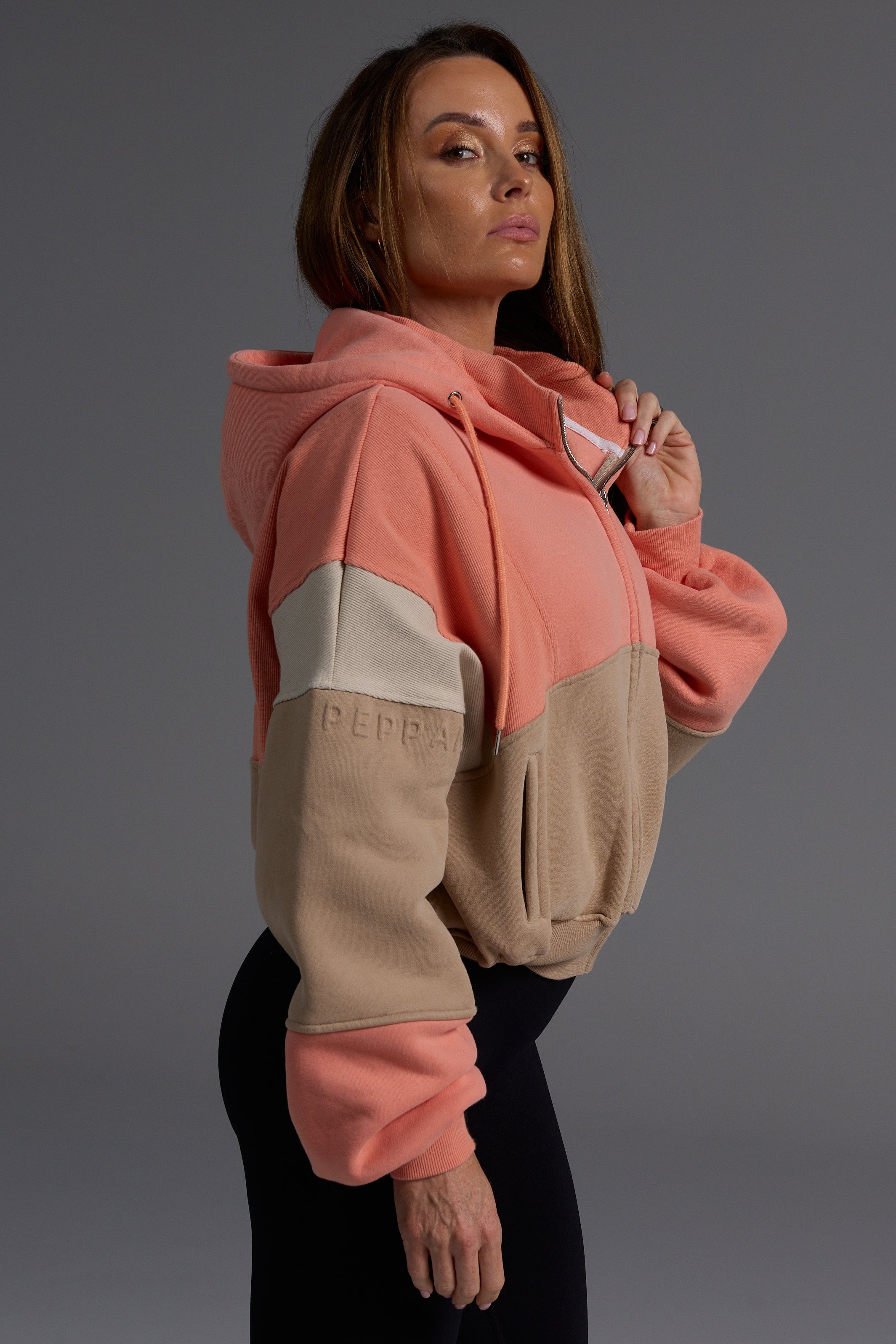 Woman wearing oversized coral and desert colorblock fleece hoodie with black leggings, showing the structured fit and premium heavyweight fabric