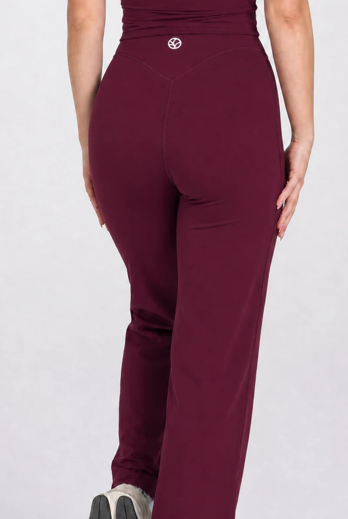 A person shown from the back wearing high-waisted burgundy wide leg pants with a small circular PEPPAMINT logo at the centre back waistband, photographed against a light grey studio background.