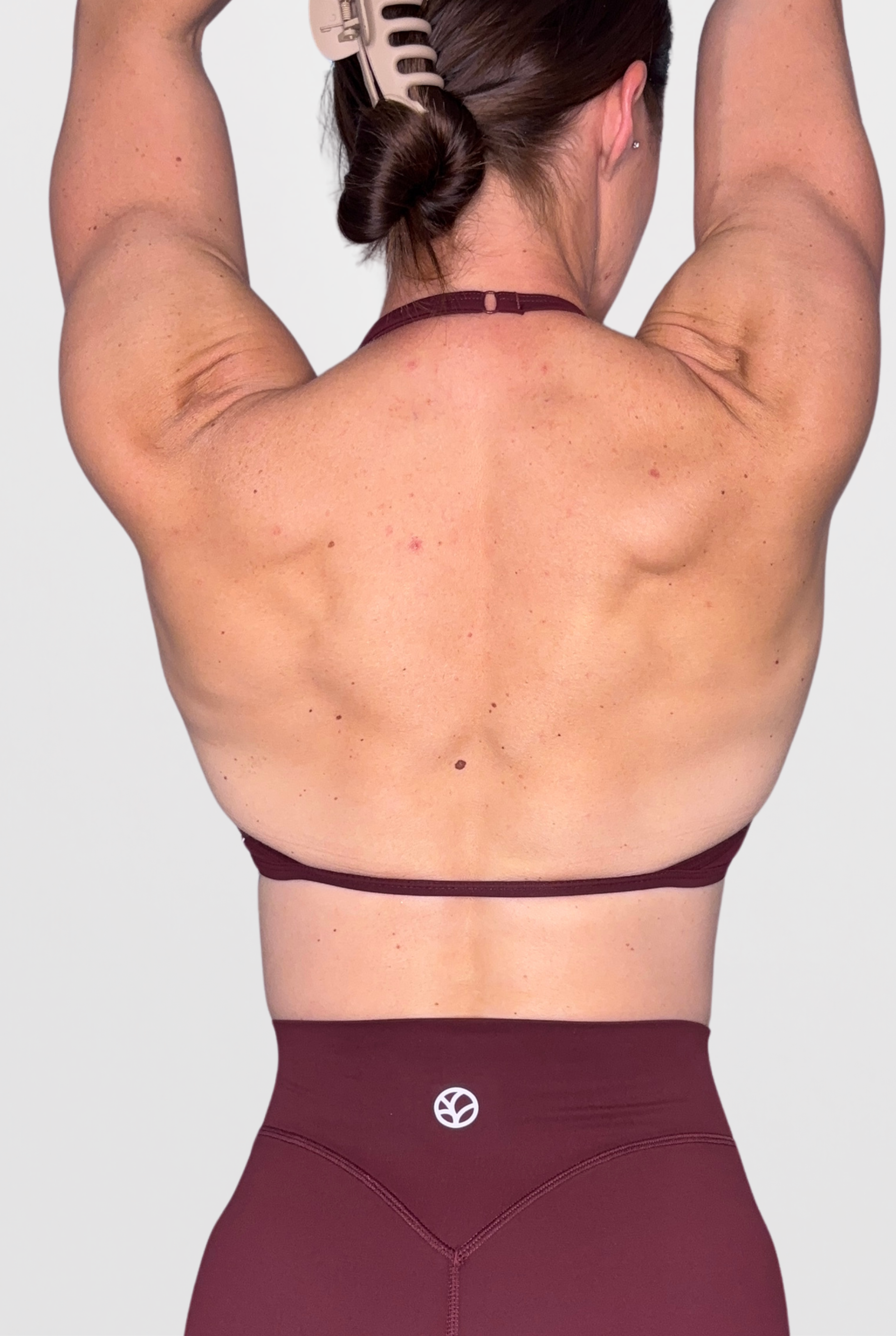 Rear view of a woman wearing a dark burgundy thin strap sports bra and matching high-waisted leggings against a light grey background.