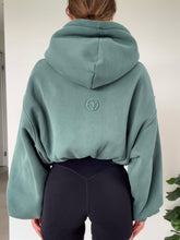 Load image into Gallery viewer, Person wearing a green hoodie and black shorts standing indoors with a plant in the background

