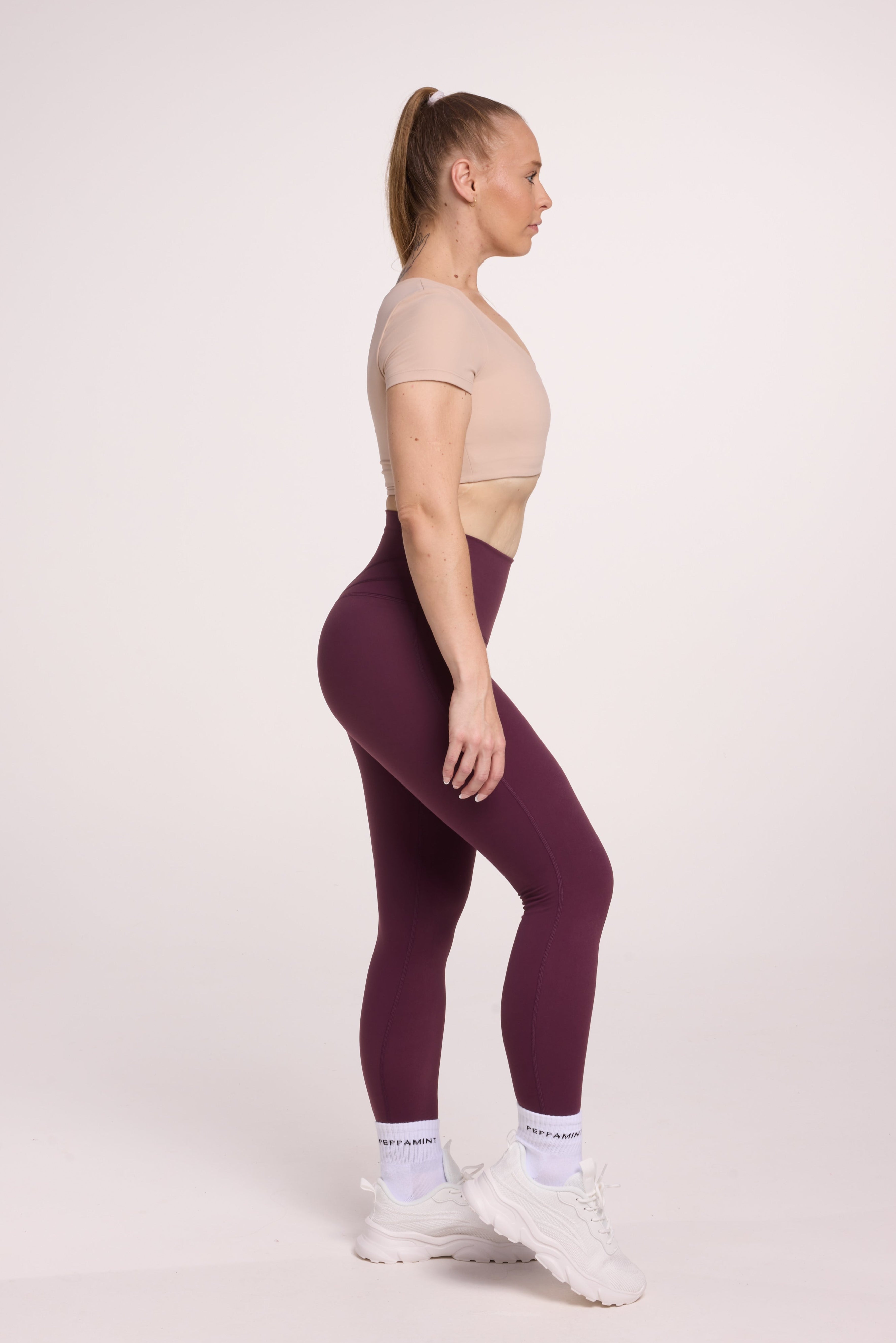 Buttery soft high waisted women's gym tights with curved back seam stitched detailing for a butt shaping effect, and no front rise seam, in plum purple paired with an V neck women's crop t-shirt with a supportive elasticated hem and triple layered fabric with removable pads for extra support in beige