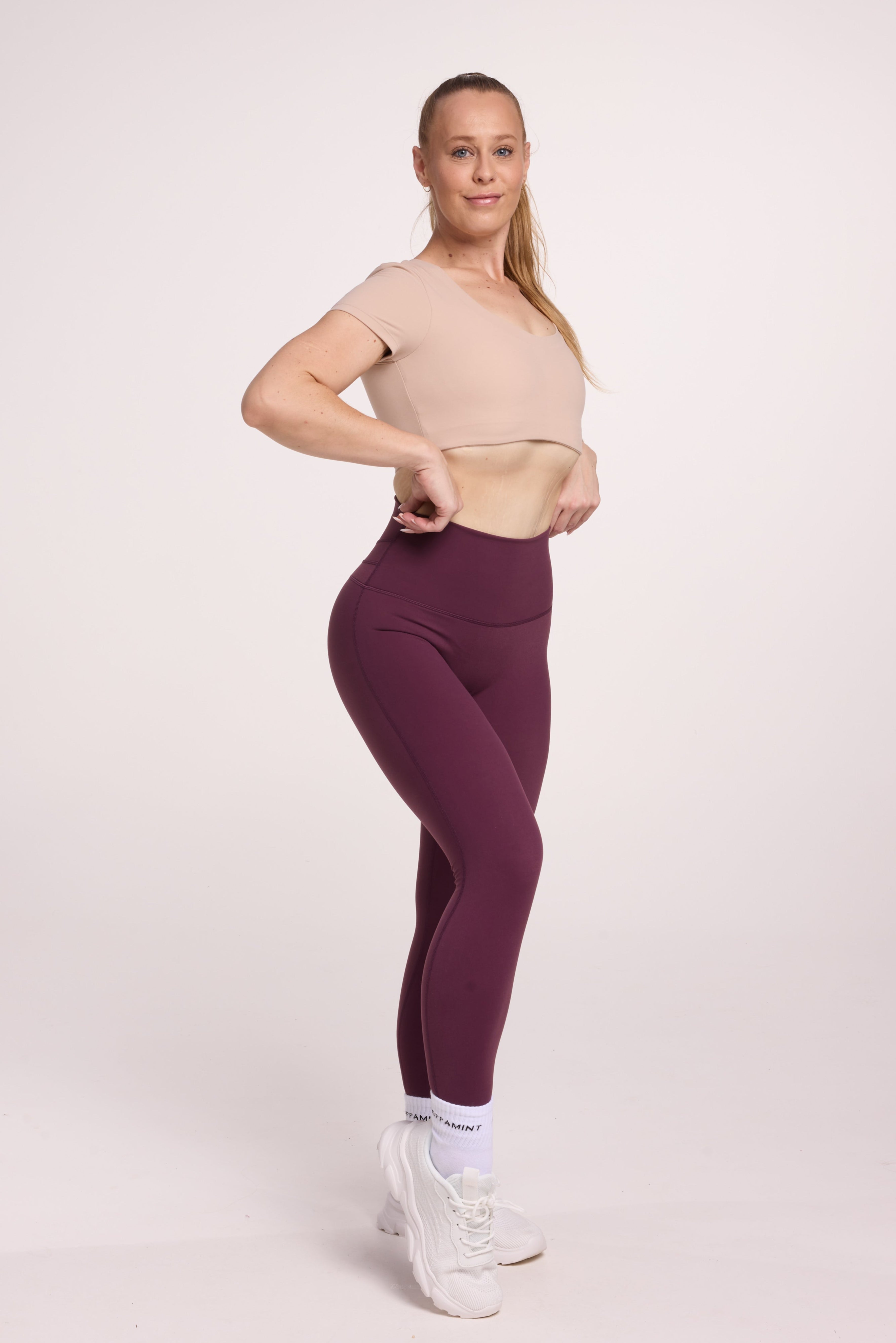 Buttery soft high waisted women's gym tights with curved back seam stitched detailing for a butt shaping effect, and no front rise seam, in plum purple paired with an V neck women's crop t-shirt with a supportive elasticated hem and triple layered fabric with removable pads for extra support in beige