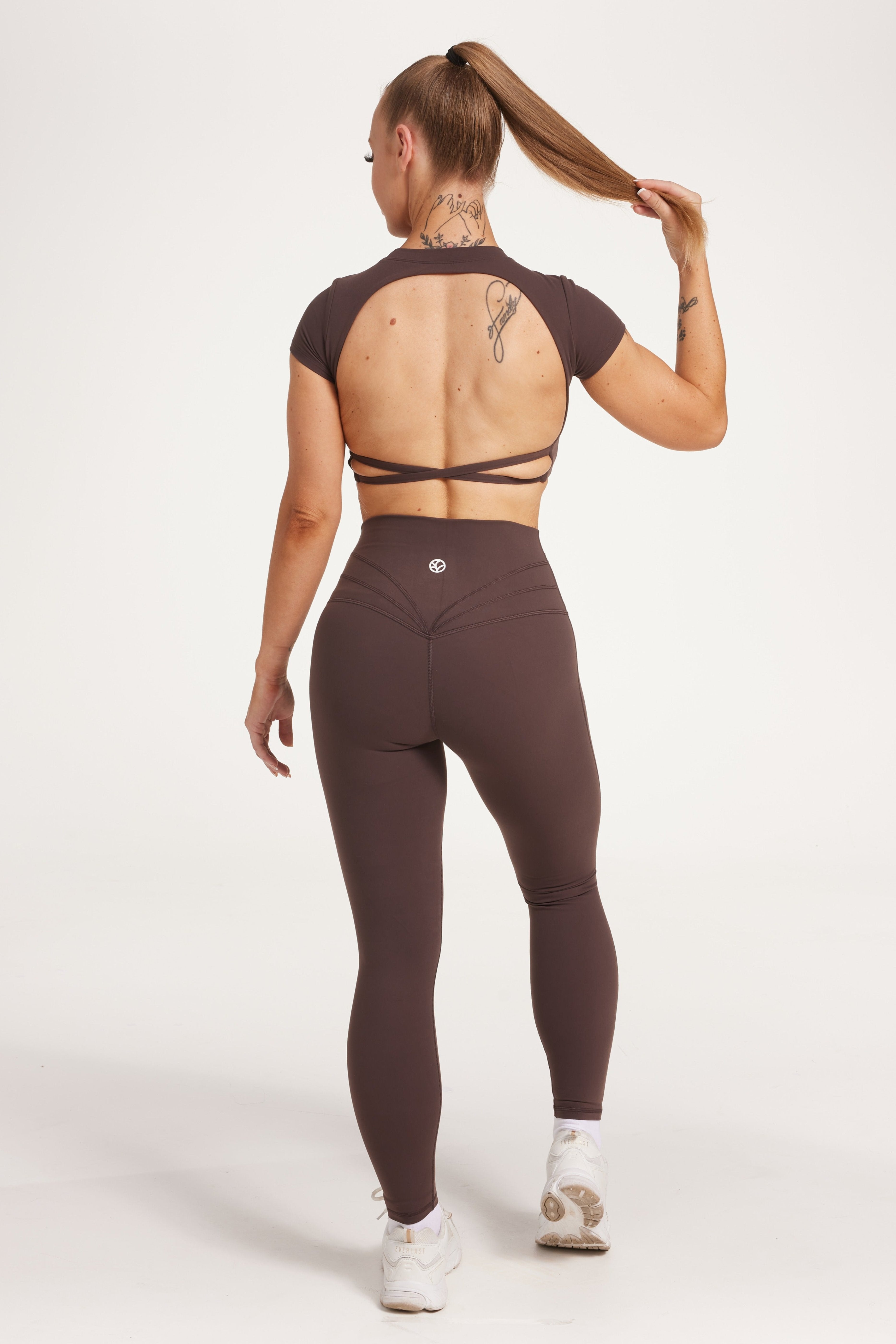 Buttery soft high waisted women's gym tights with curved hem stitched detailing for a butt shaping effect, and no front rise seam, paired with an open back women's crop t-shirt with a supportive elasticated hem and triple layered fabric with removable pads for extra support and aesthetic cross back straps in dark chocolate.