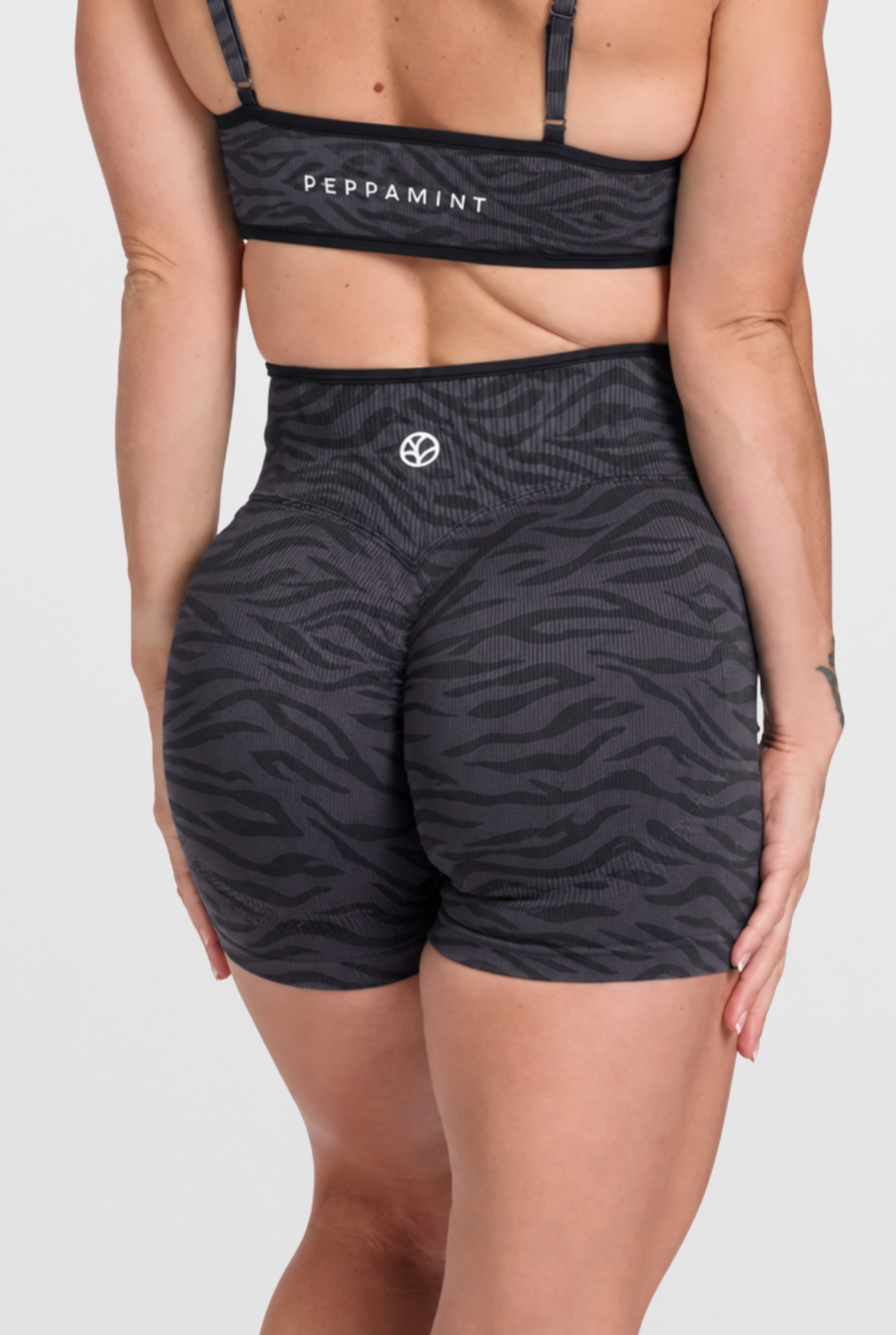 Rear view of a model wearing a matching activewear set with a dark gray and black zebra print, featuring a sports bra with 'PEPPAMINT' text and high-waisted shorts with black trim and a small white logo.