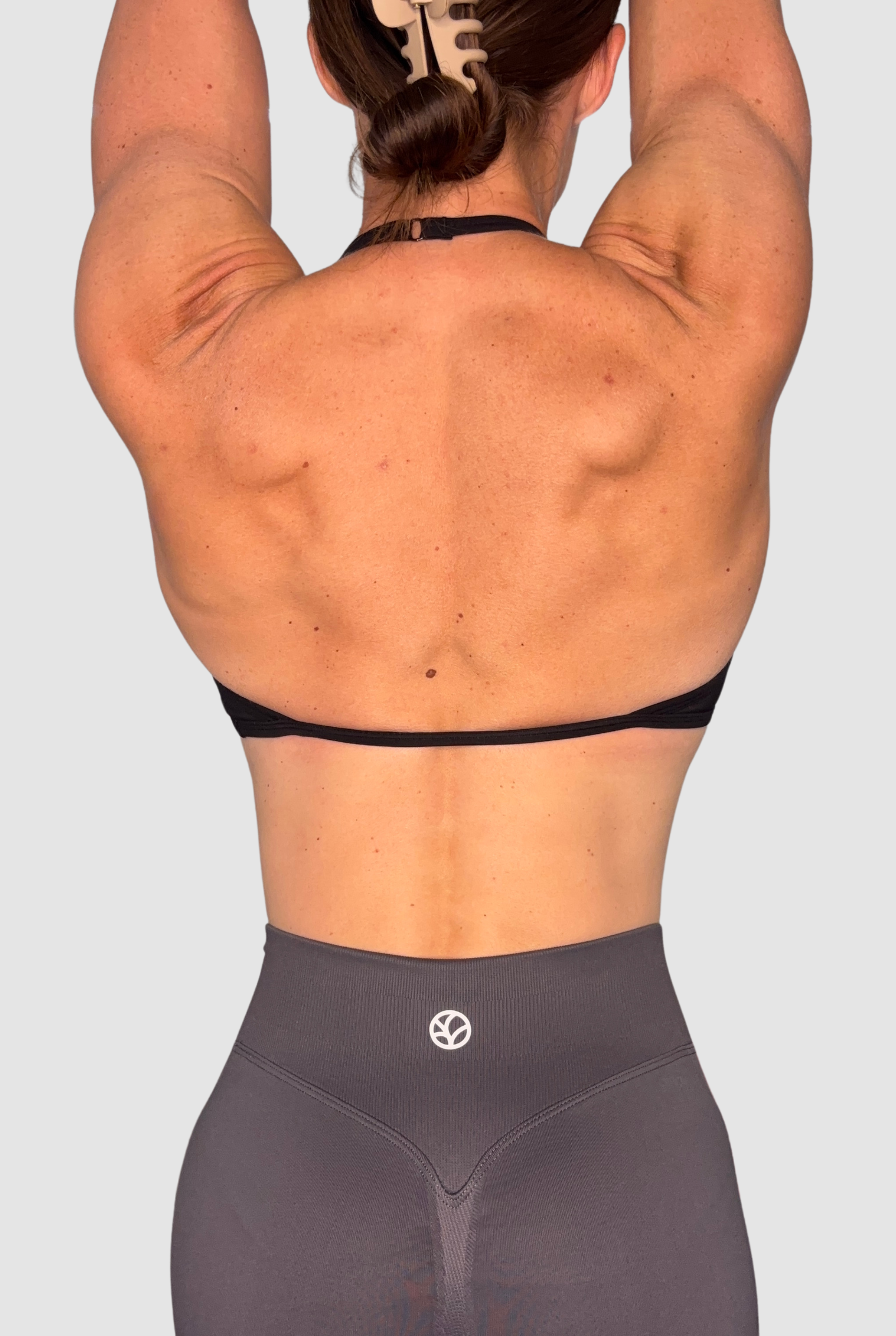 Rear view of a woman with her arms raised overhead, wearing a black sports bra and dark grey high-waisted shorts with a small white PEPPAMINT circular logo on the back waistband.