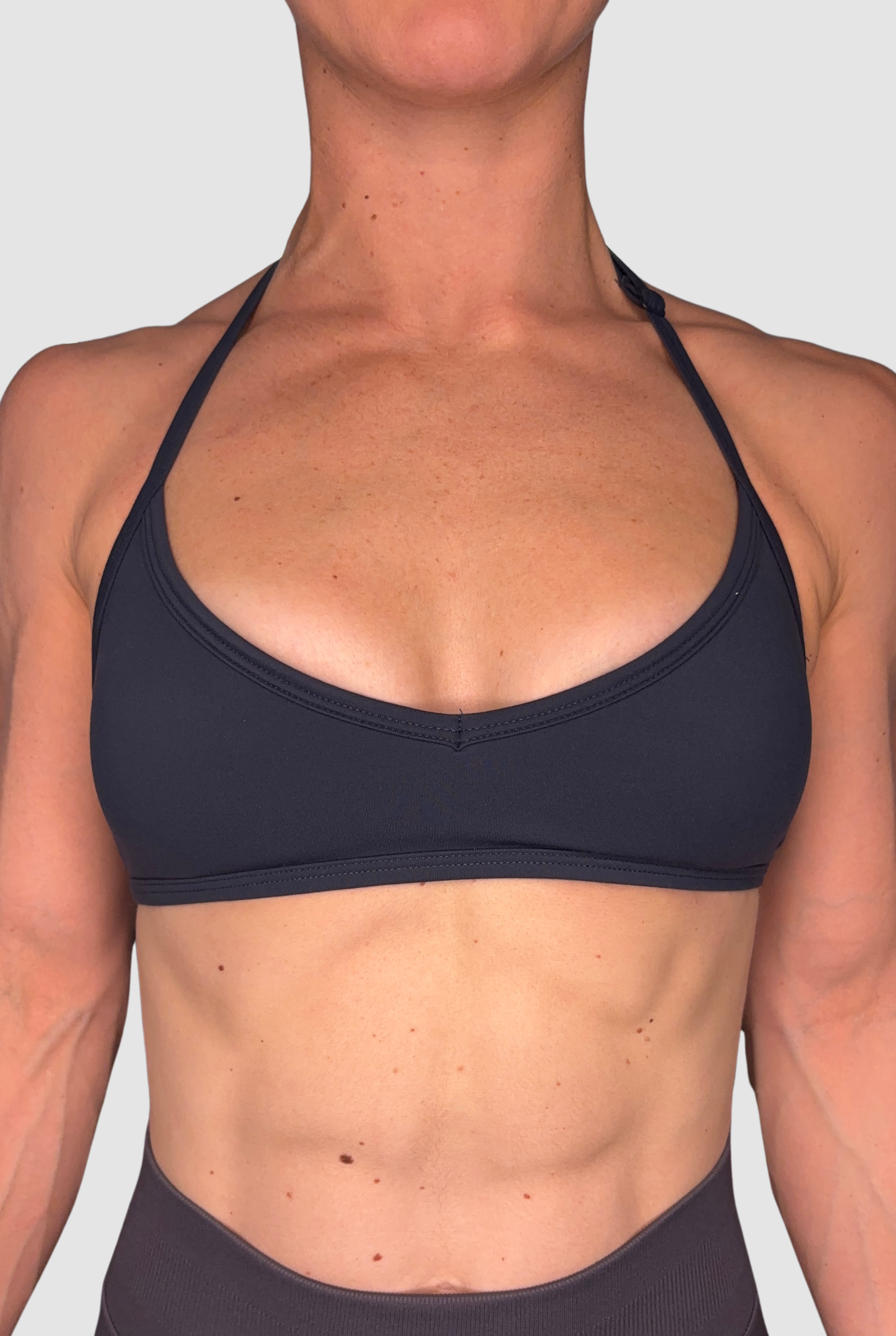 A close-up of a woman wearing a dark navy sports bra with an adjustable halter neck against a light grey background.