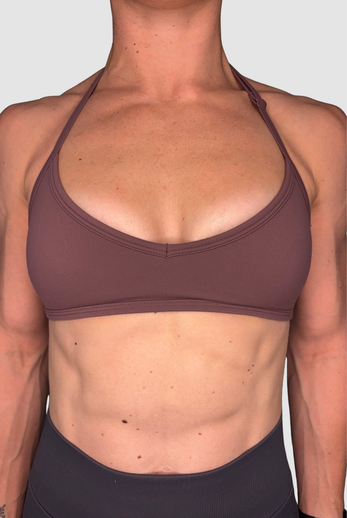 A close-up of a woman's torso wearing a muted mauve-brown sports bra with an. adjustable halter neck and dark grey athletic bottoms against a plain light grey background.