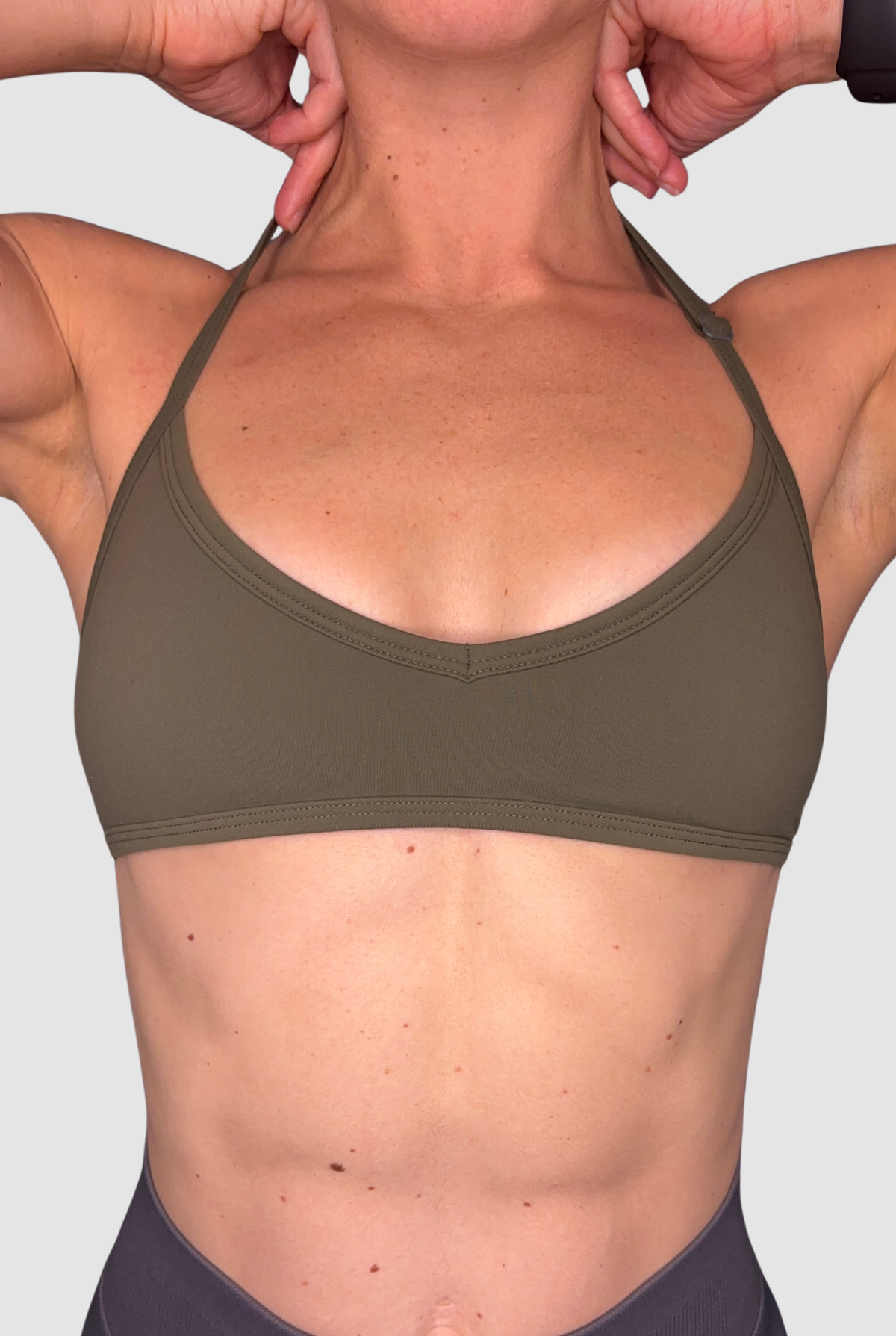 A close-up of a woman wearing an olive green sports bra, with an adjustable halter neck, showing the front of the bra and her neck, chest, and midsection against a light grey background.
