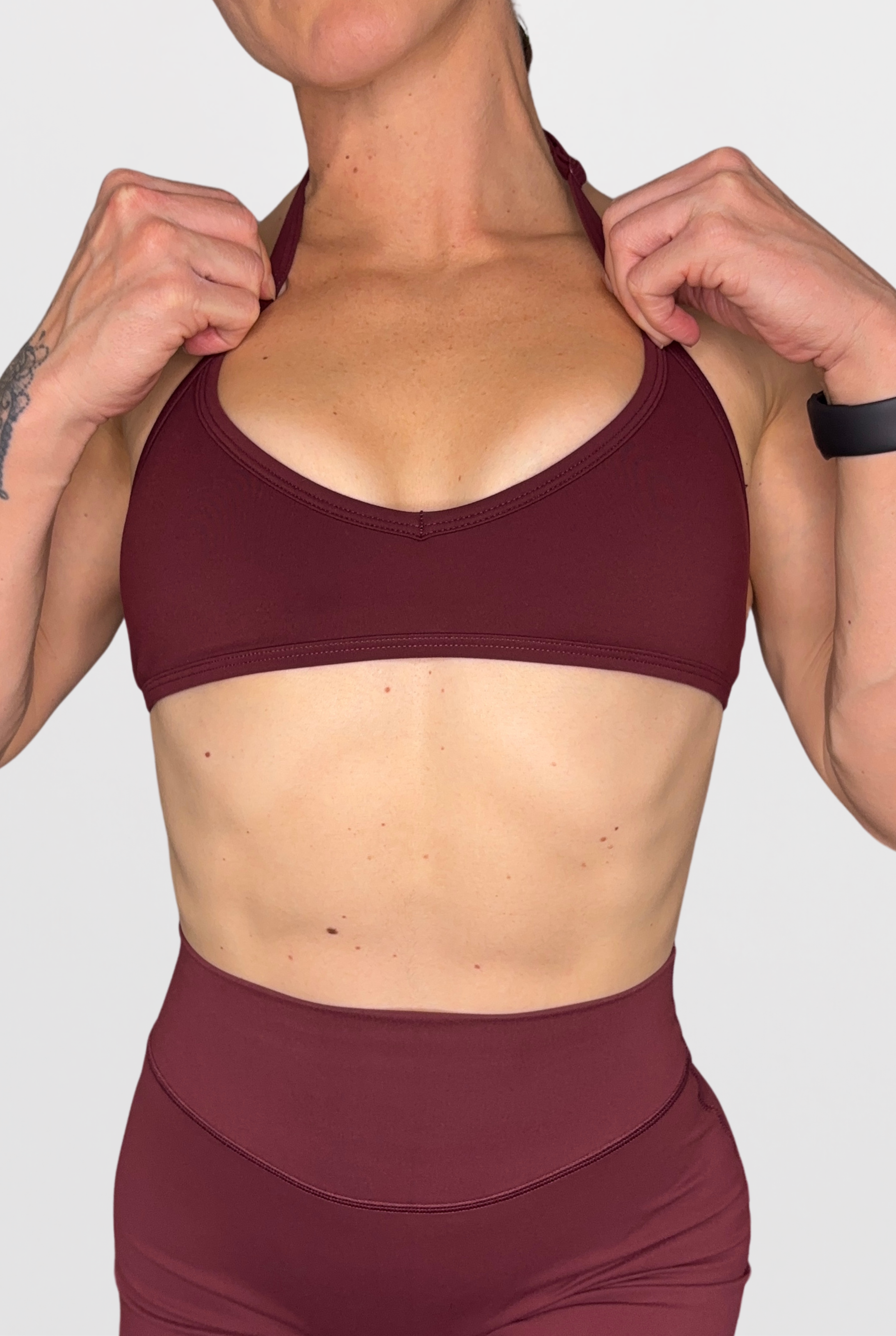 A close-up of a woman wearing a matching burgundy sports bra and high-waisted leggings, adjusting the straps of the sports bra against a light grey background.