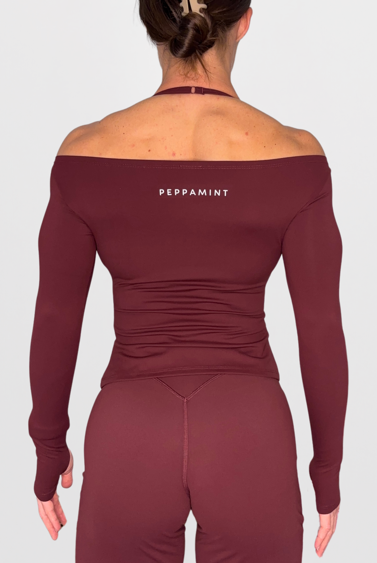 Rear view of a woman wearing a long-sleeve off-the-shoulder athletic top and matching leggings in a deep burgundy shade, with the brand name 'PEPPAMINT' printed in white on the upper back. The background is a clean, light grey studio backdrop.
