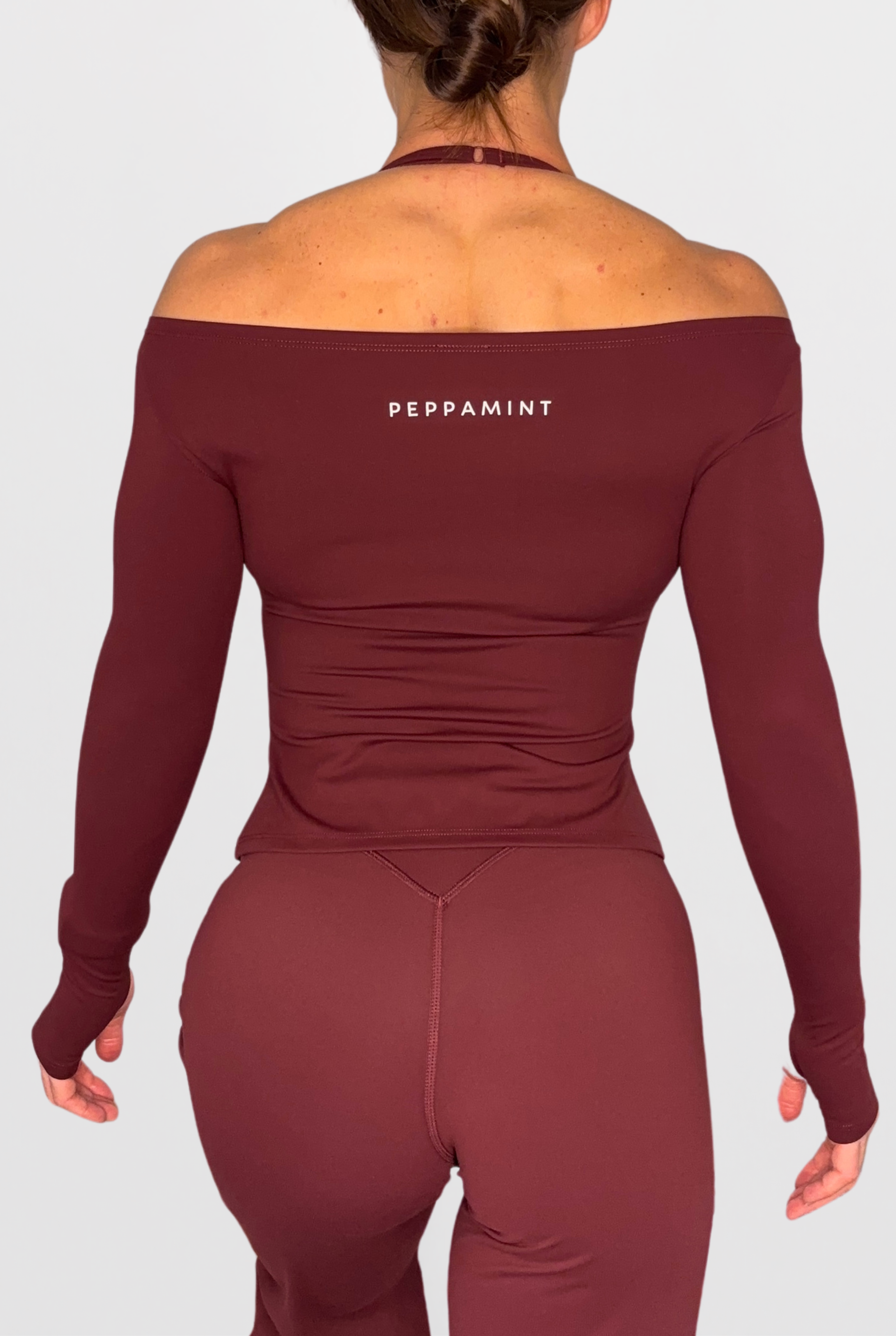 Rear view of a woman wearing a long-sleeve off-the-shoulder athletic top and matching leggings in a deep burgundy colour, with the brand name 'PEPPAMINT' printed in white on the upper back.