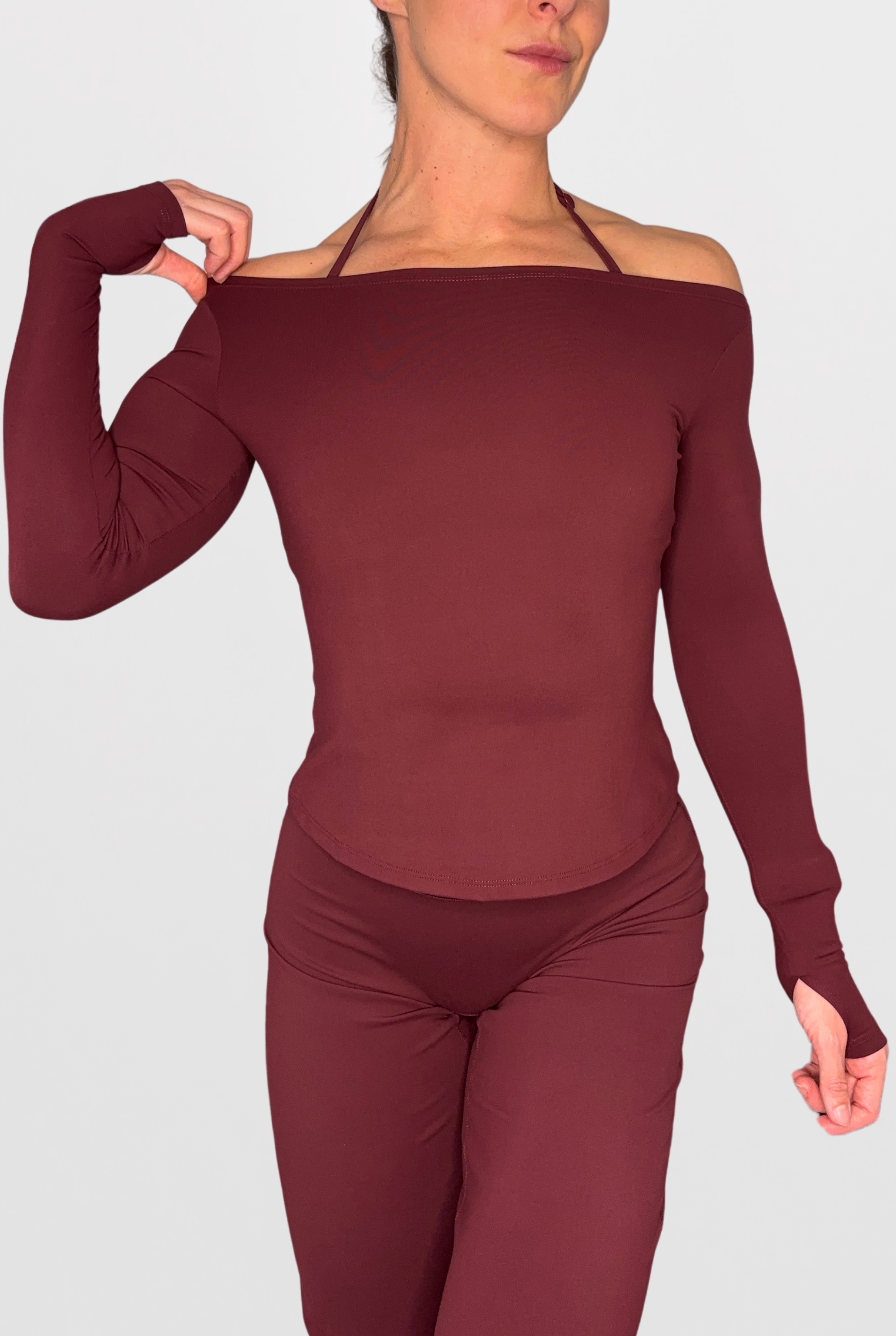 A woman modeling a matching burgundy off-the-shoulder long-sleeve top and leggings set against a light grey studio background.