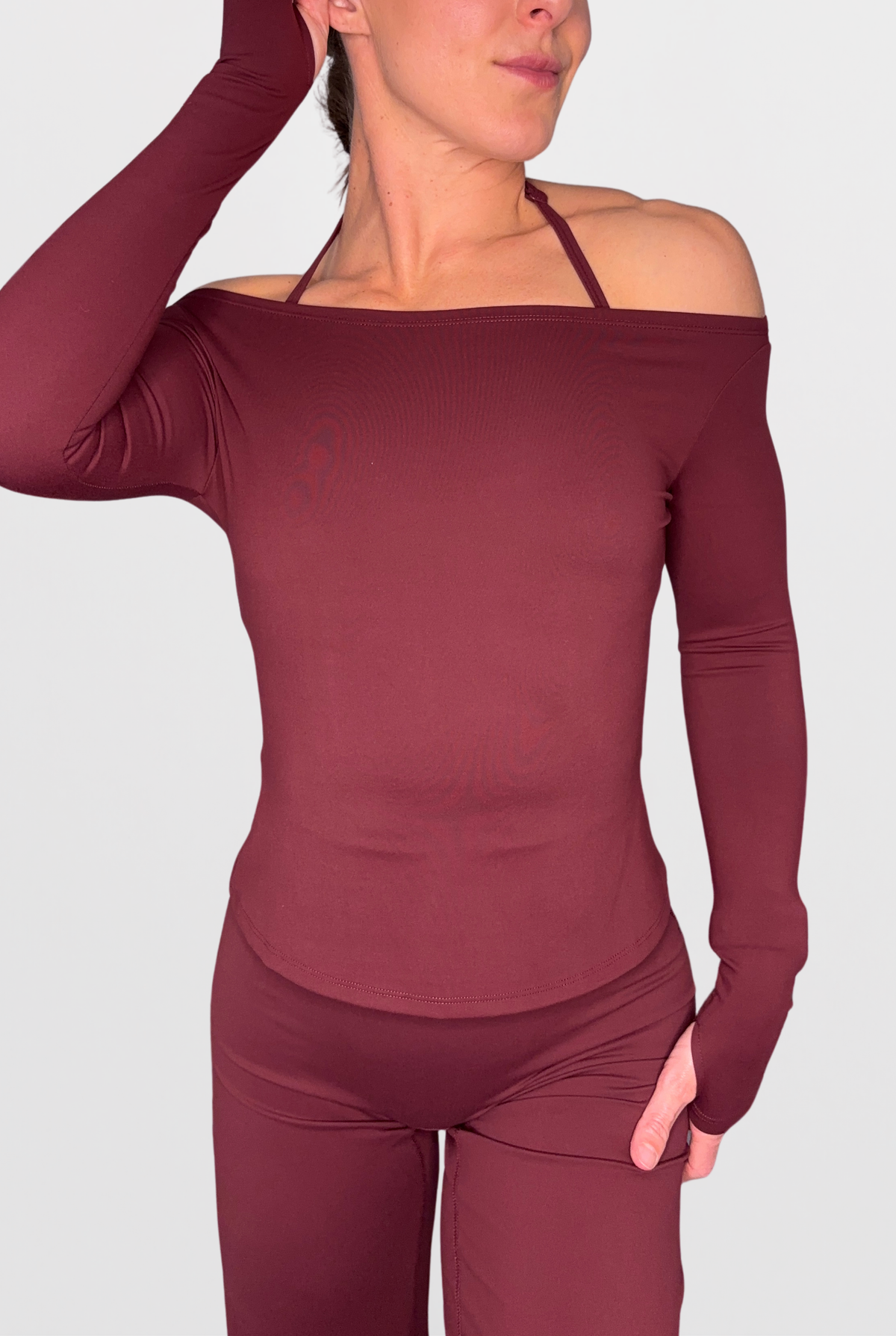 A woman wearing a long-sleeve off-the-shoulder athletic top and matching leggings in a deep burgundy colour, shown against a solid light grey background.