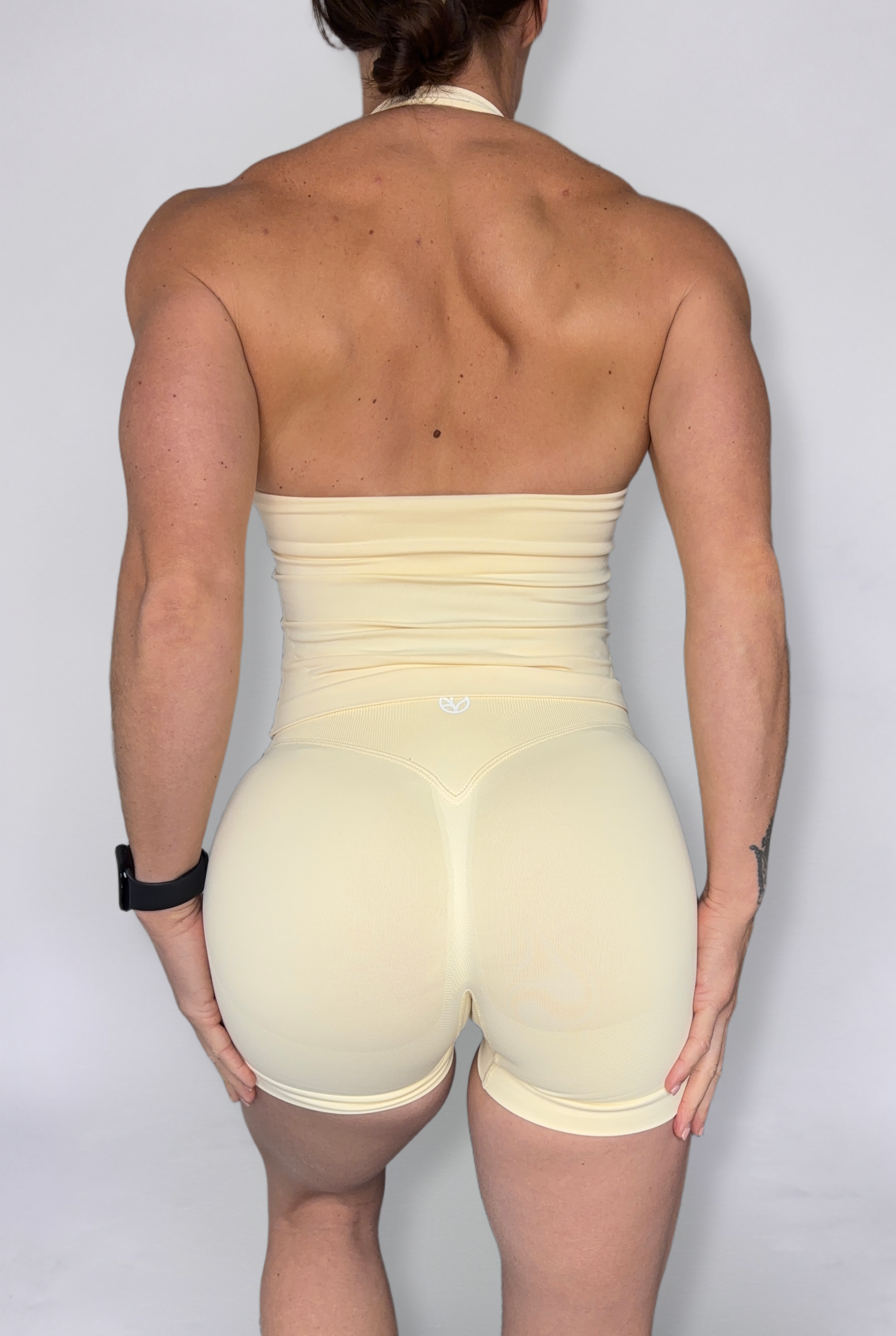 Rear view of a woman wearing a pale yellow high-waisted shorts and open back halter tank, set against a plain light gray background.